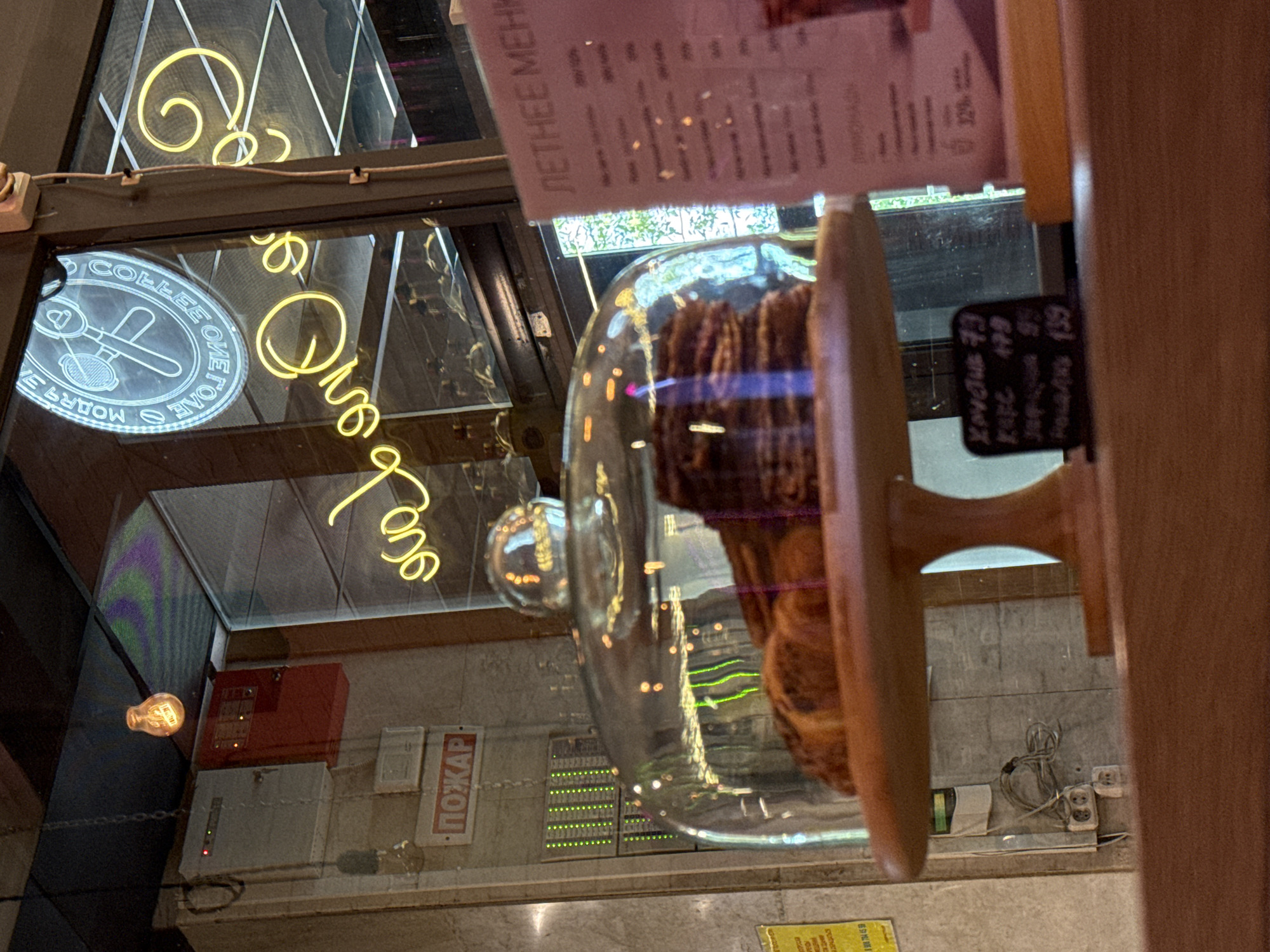 The image depicts a dessert display case at a cafe or bakery, likely in Moscow, Russia. A stack of pastries, possibly croissants, is visible under a glass dome on a wooden stand. In the background, a neon sign with Cyrillic script, reading "ONE COFFEE", is illuminated. To the left of the sign, a fire alarm panel is mounted on a wall, with a "POZHAR" (Fire) sign above it. A menu board, with Cyrillic text, is partially visible on the right side of the image. The overall lighting suggests an indoor setting, possibly during the day, with reflections from the neon sign and lights on the glass dome.