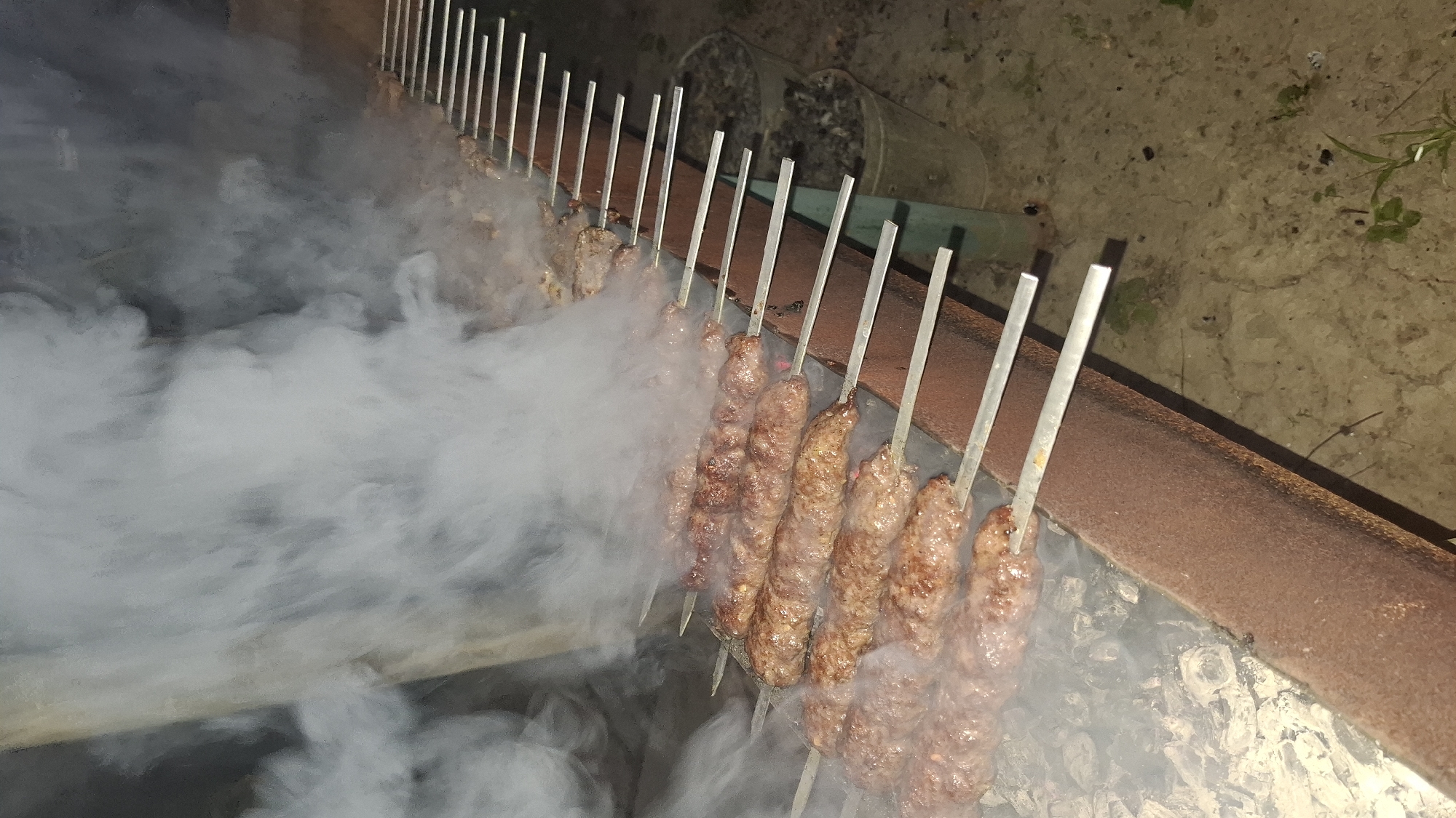 A close-up, high-angle shot captures multiple skewers of meat cooking over an open fire, likely in an outdoor setting in Шұбарсу, Kazakhstan. The meat, appearing to be ground meat formed into oblong shapes, is brown and slightly charred, indicating it is well-cooked or actively grilling. Wisps of white smoke rise from the skewers and the hot coals below, obscuring some of the background. The skewers are laid across a rusty metal grill structure. In the background, two dark buckets filled with what appear to be ashes or debris are visible. The ground beneath the grill appears to be dirt or gravel, with some sparse vegetation. The lighting suggests it may be evening or night, with the cooking fire providing the primary light source. There are no people visible in the frame.