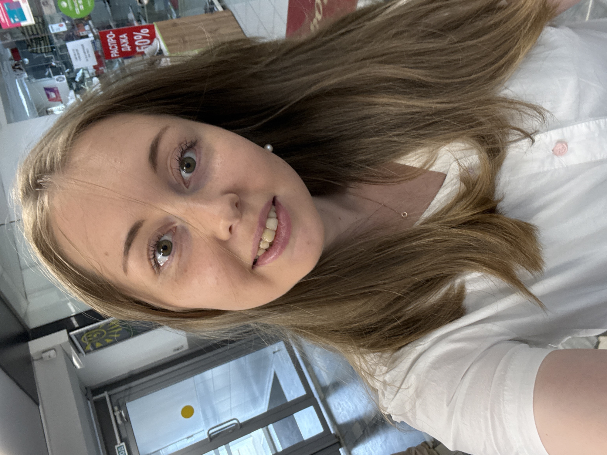 A close-up, selfie-style photograph captures a young woman with long, wavy brown hair and fair skin. She is wearing a white collared shirt with a small pink button at the collarbone and a delicate rose gold necklace. A small pearl earring dangles from her right ear. Her eyes are a light green, and she is looking directly at the camera with a slight smile. The background appears to be an indoor commercial space, possibly a store, with glass doors and shelves visible, along with a red sign displaying "РАСПРОДАЖА 50%" which translates to "SALE 50%". The lighting suggests an indoor environment, possibly with fluorescent or overhead lighting.