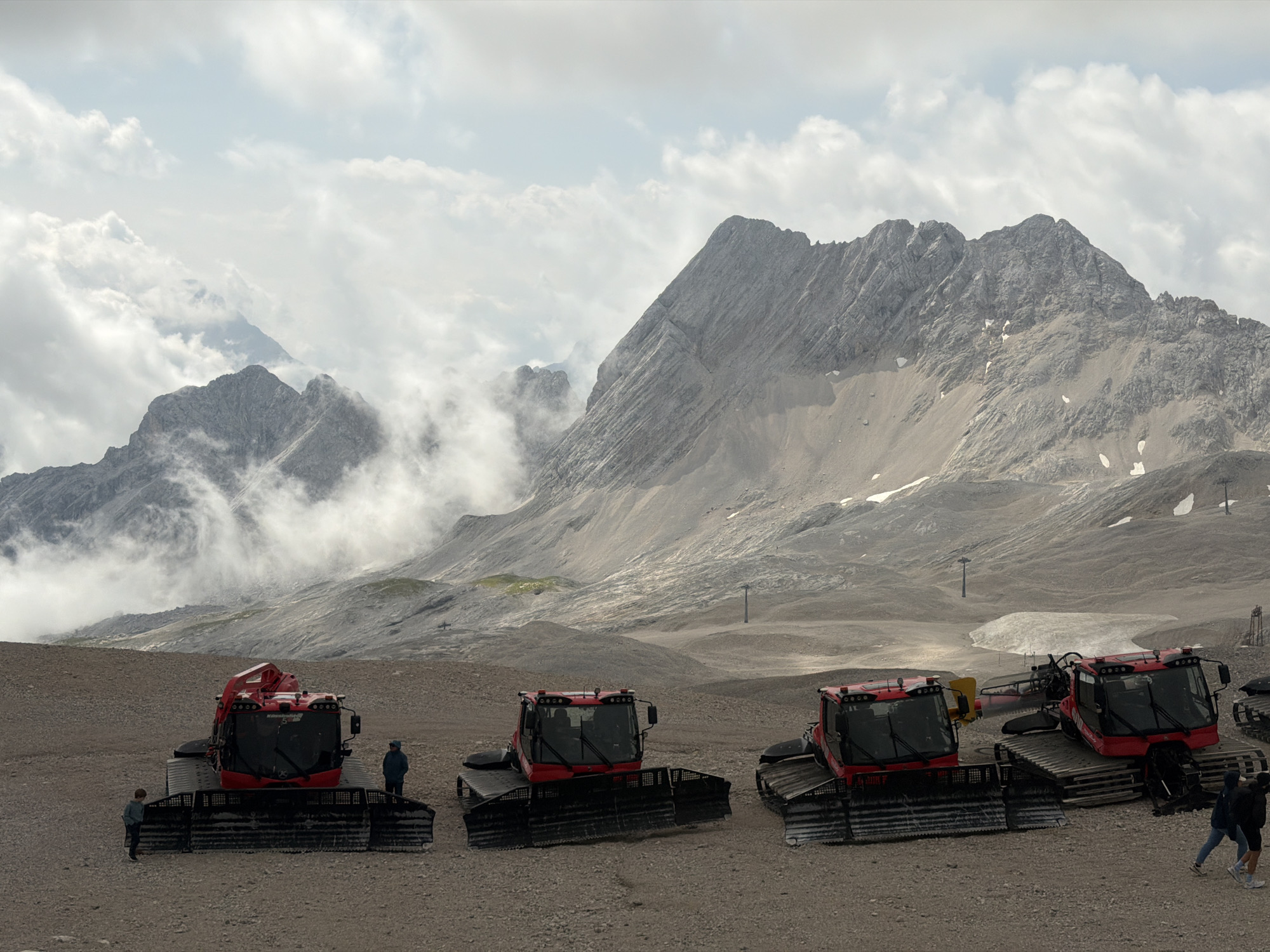 The image depicts a mountain landscape in Garmisch-Partenkirchen, Germany, likely on a high-altitude plateau. In the foreground, four red snow groomers are parked on a gravelly, uneven surface. The closest snow groomer, on the left, is red with black tracks and a wide blade. To its right, another red snow groomer is parked slightly behind and to the right. Further to the right, a third red snow groomer is positioned, and to its right, a fourth snow groomer is partially visible. Two children are standing near the first snow groomer on the left. A person in a blue jacket stands to the right of the first snow groomer. Two individuals, one in a dark jacket and the other in a blue jacket, are walking away from the viewer on the far right of the frame. In the middle ground, the terrain is barren and rocky, with patches of snow. In the background, imposing, rugged mountains rise under a cloudy sky. The mountains are characterized by steep, rocky slopes, with some areas showing signs of erosion and snow accumulation. Wisps of clouds cling to the mountain peaks and drift across the sky, partially obscuring some of the distant formations. The overall scene conveys a sense of a remote, high-altitude environment.