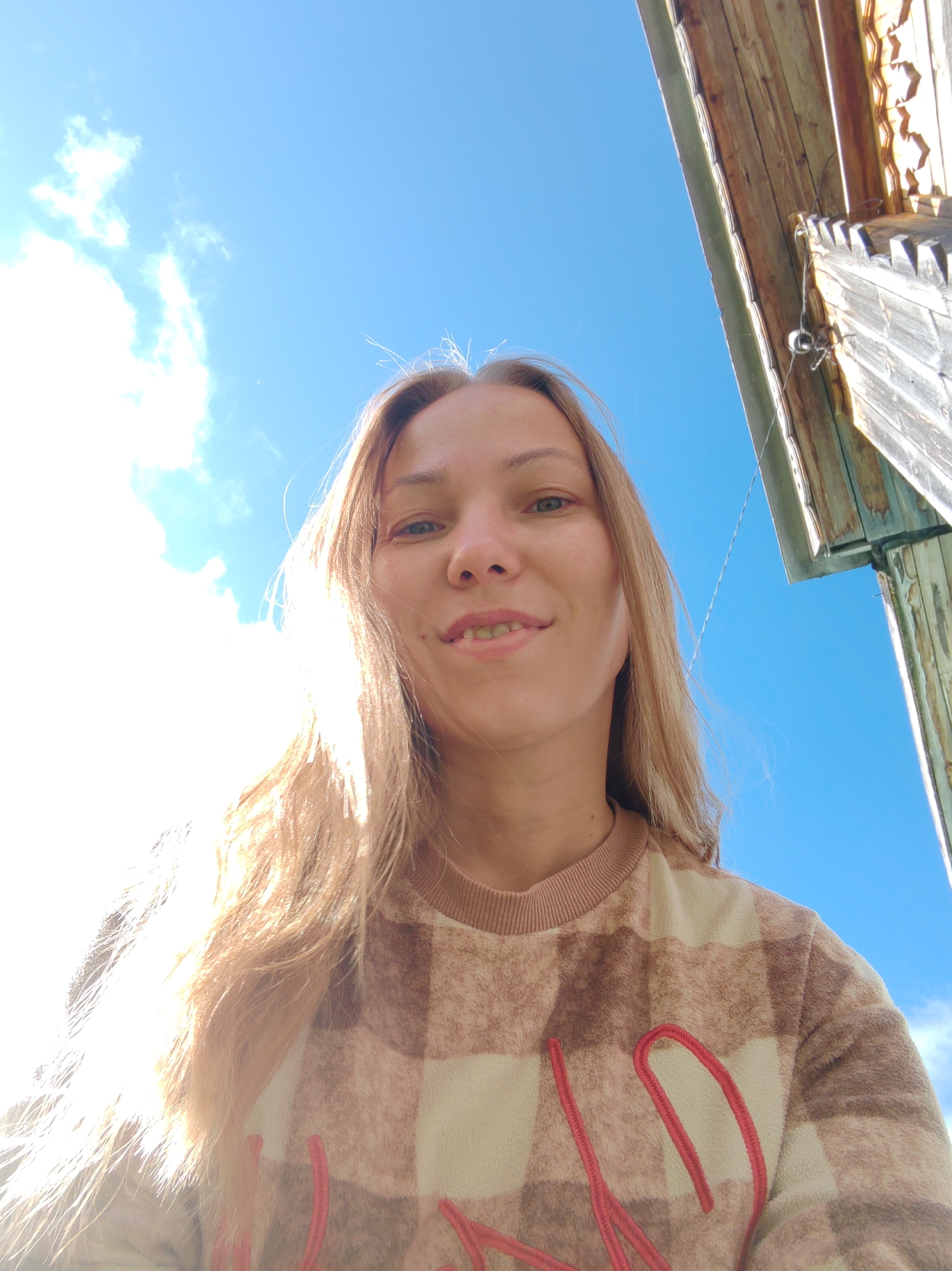 A low-angle, outdoor selfie shows a Caucasian woman with long blonde hair and light skin. She is smiling and looking at the camera. She is wearing a beige and brown plaid fleece sweatshirt with red lettering on the front. The background consists of a bright blue sky with a few white clouds, and the wooden roof and eaves of a building are visible in the upper right corner. The image appears to have been taken on a sunny day. The building's construction suggests it may be in a rural or older part of the city of Chukhlom, Russia.