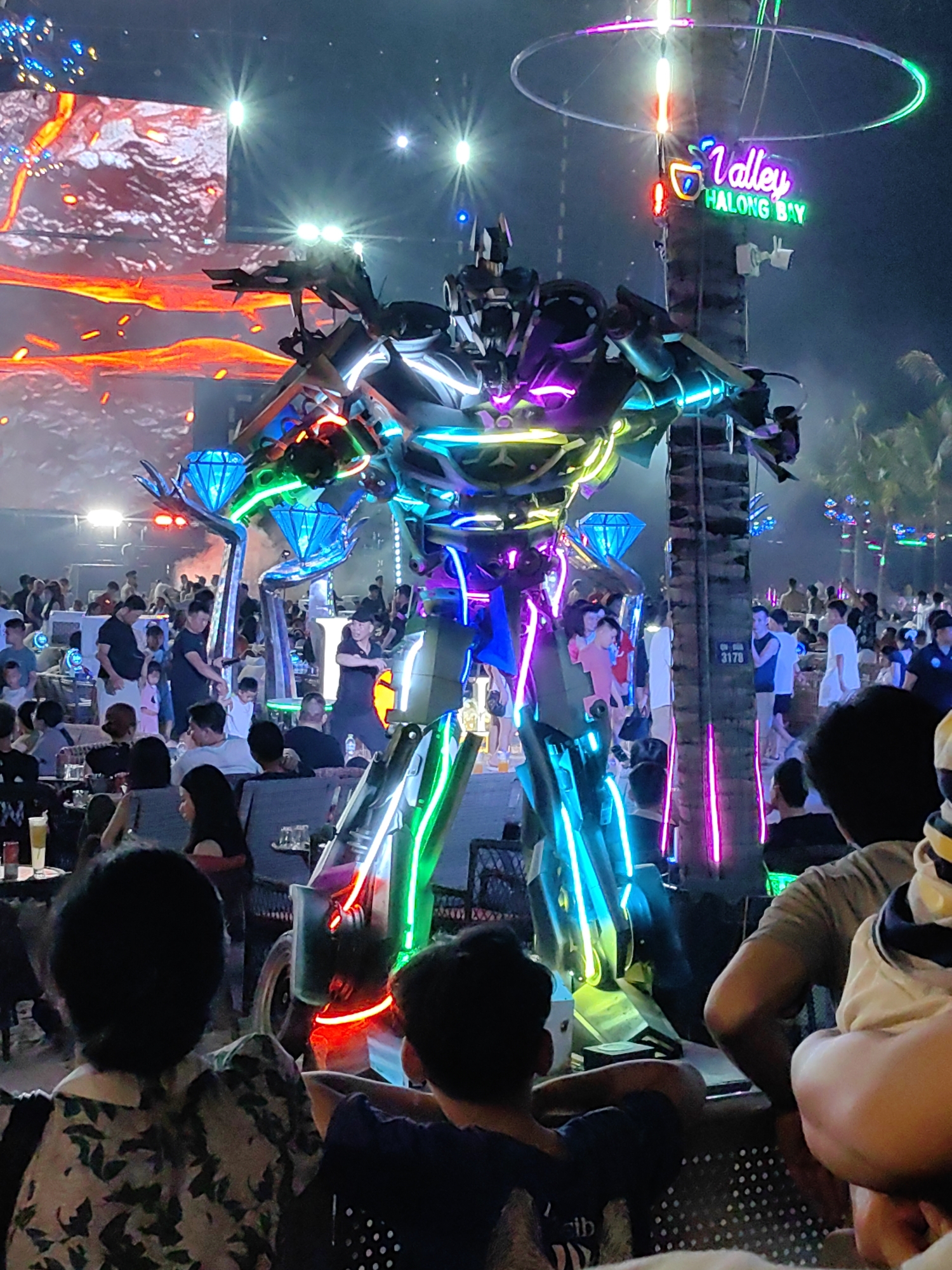 A nighttime scene in Bai Chay Ward, Vietnam, shows a large, brightly lit robotic figure standing among a crowd of people. The robot, adorned with neon lights of various colors (green, blue, purple, and yellow), appears to be the center of attention. People are seated at tables and standing around, observing the spectacle. In the background, a video screen displays images of lava or fire. A neon sign reading "Valley Halong Bay" is visible, attached to a palm tree. The scene suggests a lively outdoor event or performance.