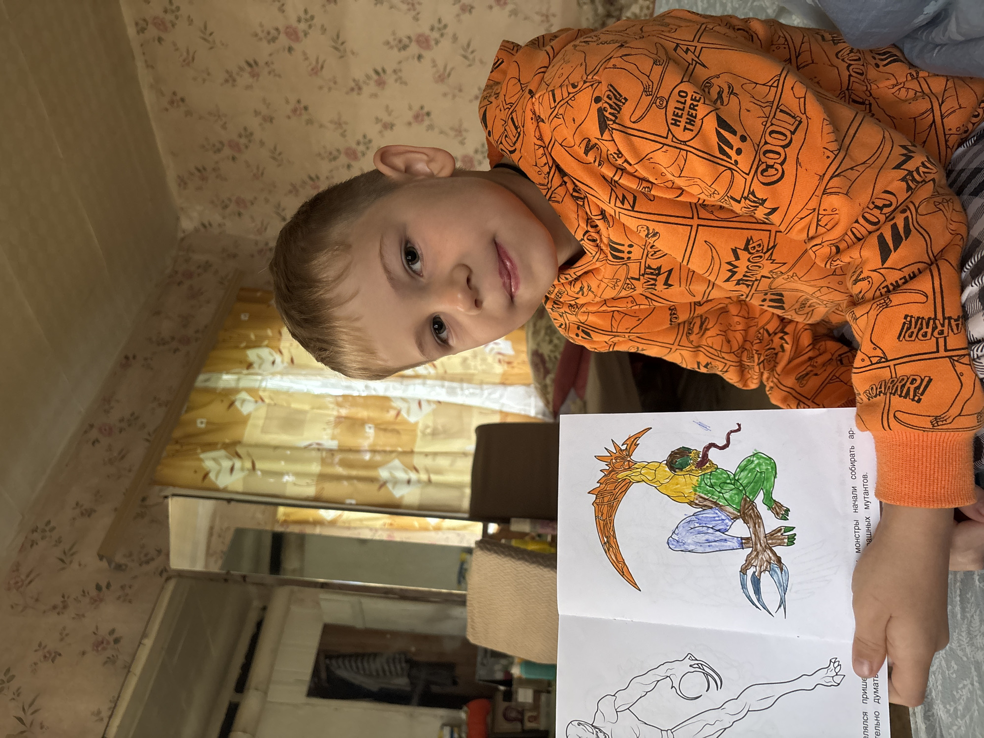 A young boy with blond hair and a light complexion is sitting on a bed in a room. He is wearing an orange hoodie with black dinosaur illustrations. He is holding an open coloring book, showing a partially colored drawing of a mythical creature with claws and a long tongue. The room has floral wallpaper, a mirror reflecting the room, and yellow curtains. The scene appears to be in a residential setting, possibly in Cherkasovo, Russia.