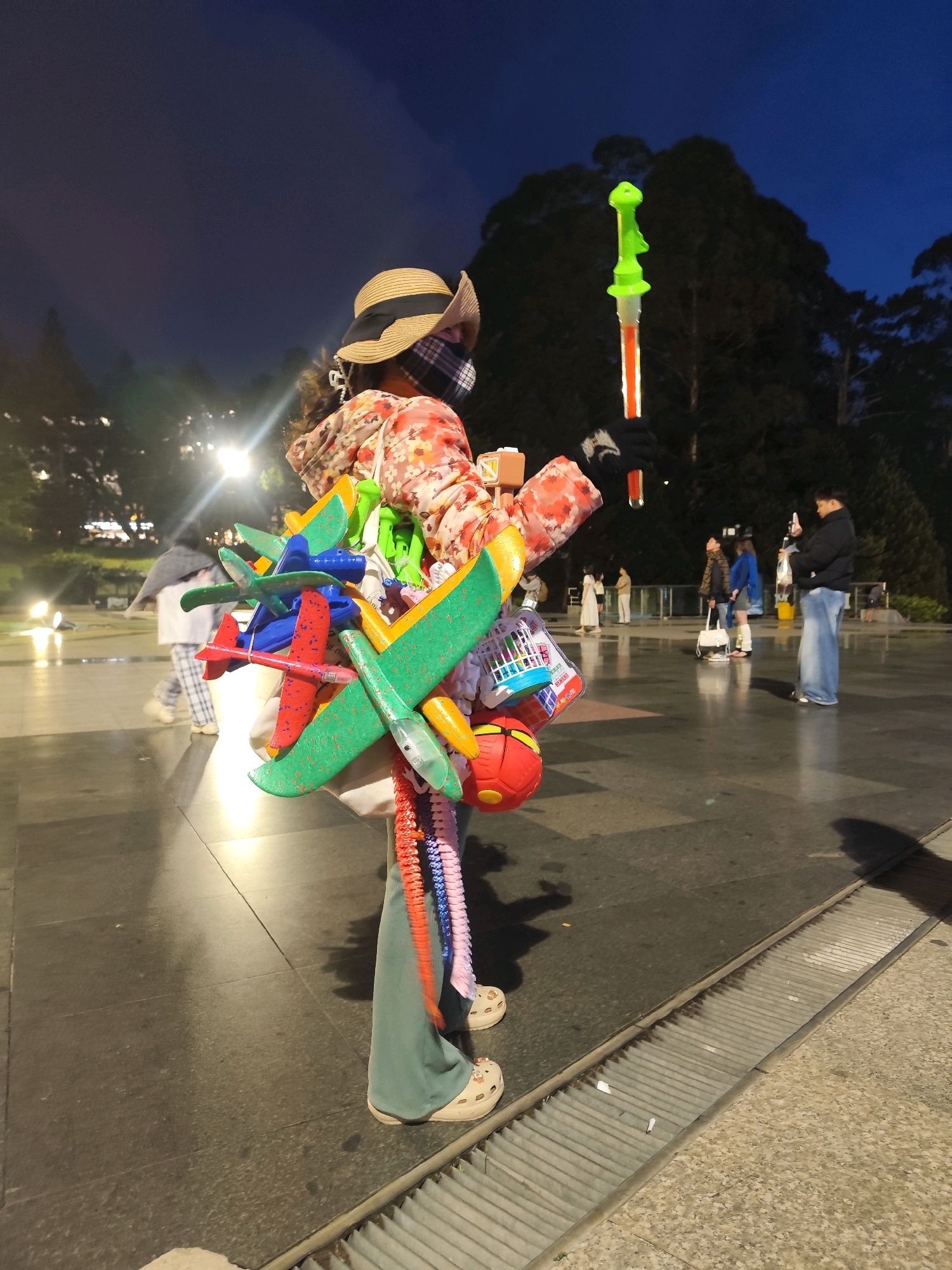 A person wearing a straw hat, a floral shirt, and wide-leg pants stands in a paved urban area at dusk. They are carrying a large collection of colorful toy airplanes and a green bubble wand with a light. The person is facing away from the camera, and a checkered face mask is visible. In the background, several other people are scattered across the plaza, some looking at their phones, and others walking. The environment features trees and streetlights, suggesting an outdoor public space. The ground is wet, reflecting the ambient light. The scene appears to be located in the vicinity of the Da Lat Night Market in Da Lat, Vietnam, a popular tourist destination known for its vibrant atmosphere and evening activities.