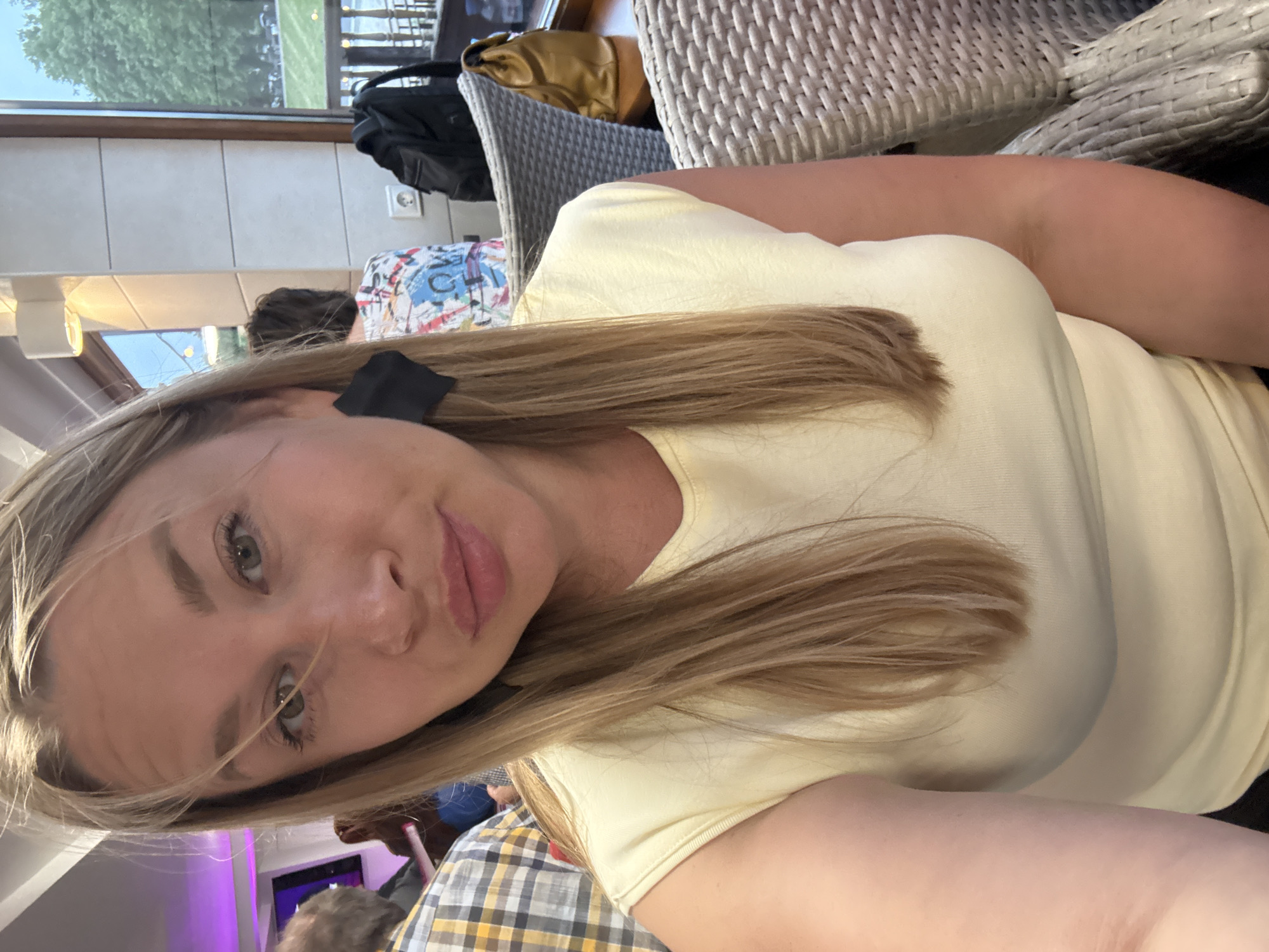 The media file is a selfie taken by a young woman with blonde hair and green eyes. She is wearing a light yellow t-shirt and a plaid shirt. Her hair is long and wavy, and she has a black hair accessory clipped to the side of her head. The background shows a dining area with wicker chairs and a table. There is a window with trees and what appears to be a street outside. Other people are visible in the background, one of whom is wearing a colorful shirt. The lighting suggests it is daytime, possibly indoors in a restaurant or cafe. The overall mood seems relaxed and casual. The location is identified as Veliky Novgorod, Russia, though there are no specific visual cues in the image to confirm this.