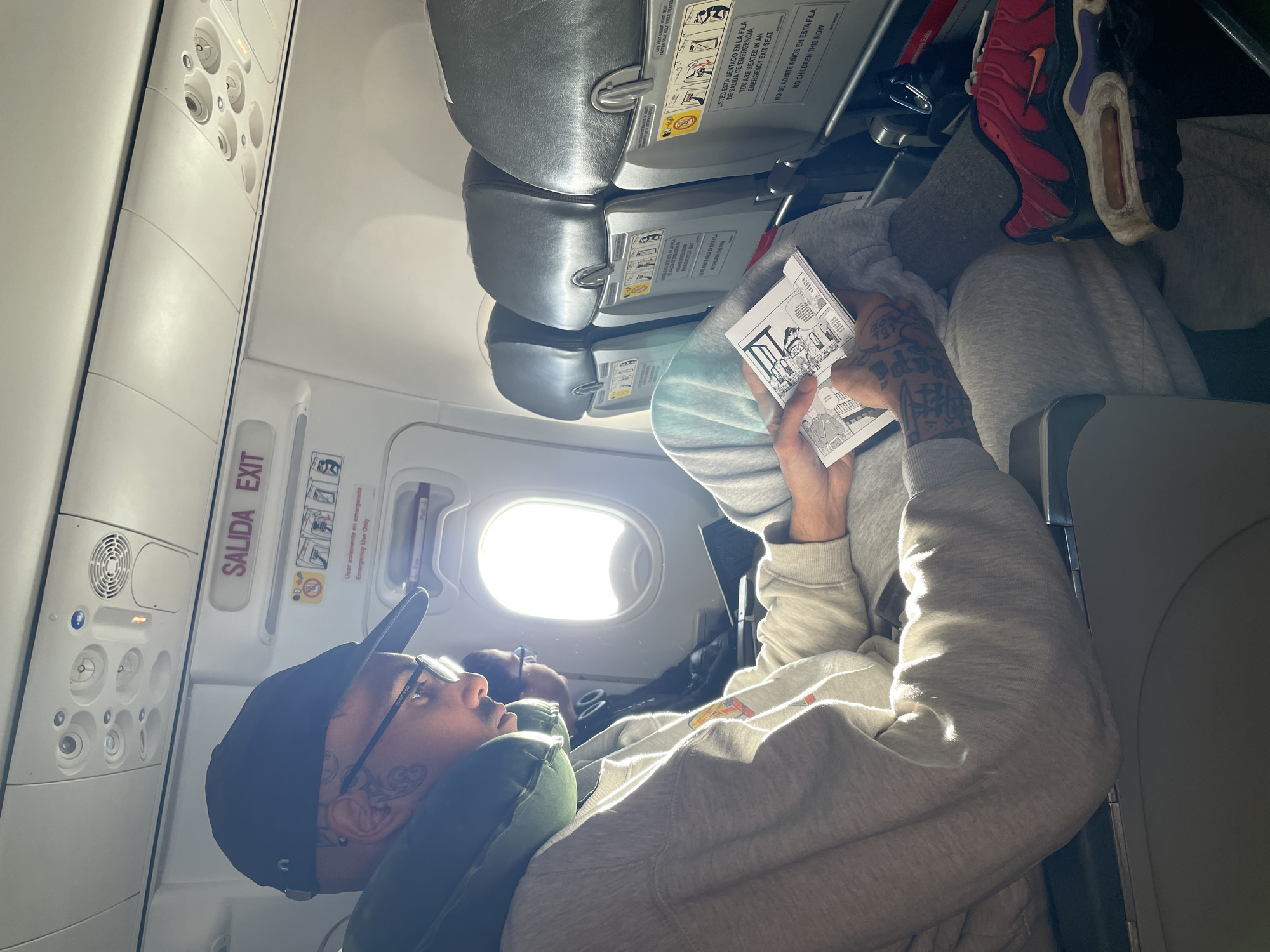 A person is captured sitting in an airplane window seat, likely at Aeropuerto Internacional Ezeiza, Argentina, given the bilingual signage. The main subject, appearing to be male, is seen wearing a black baseball cap, glasses, a grey hoodie, and grey sweatpants. Distinctive tattoos adorn his hands and neck/face. An inflatable green neck pillow rests around his neck, and red and purple Nike Air Max sneakers are visible on his propped-up foot. He holds a small comic book or booklet, appearing relaxed as he reads or looks forward/out the brightly lit window. A faint reflection of another person's head is visible in the window.
Bright daylight floods the cabin through the window, indicating it is daytime. The overhead panel with reading lights and air vents is visible above. Other grey airplane seats are in front of him. The emergency exit door is prominent with clear signage.
Visible text includes: "SALIDA EXIT," "Usar solamente en emergencia / Emergency Use Only," "LIFE VEST UNDER YOUR SEAT / WHILE SEATED," and specific instructions for an emergency exit row: "USTED ESTA SENTADO EN LA FILA DE SALIDA DE EMERGENCIA / YOU ARE SEATED IN AN EMERGENCY EXIT SEAT" and "NO SE ADMITE NIÑOS EN ESTA FILA / NO CHILDREN THIS ROW." The number "8" is also visible on the overhead console.