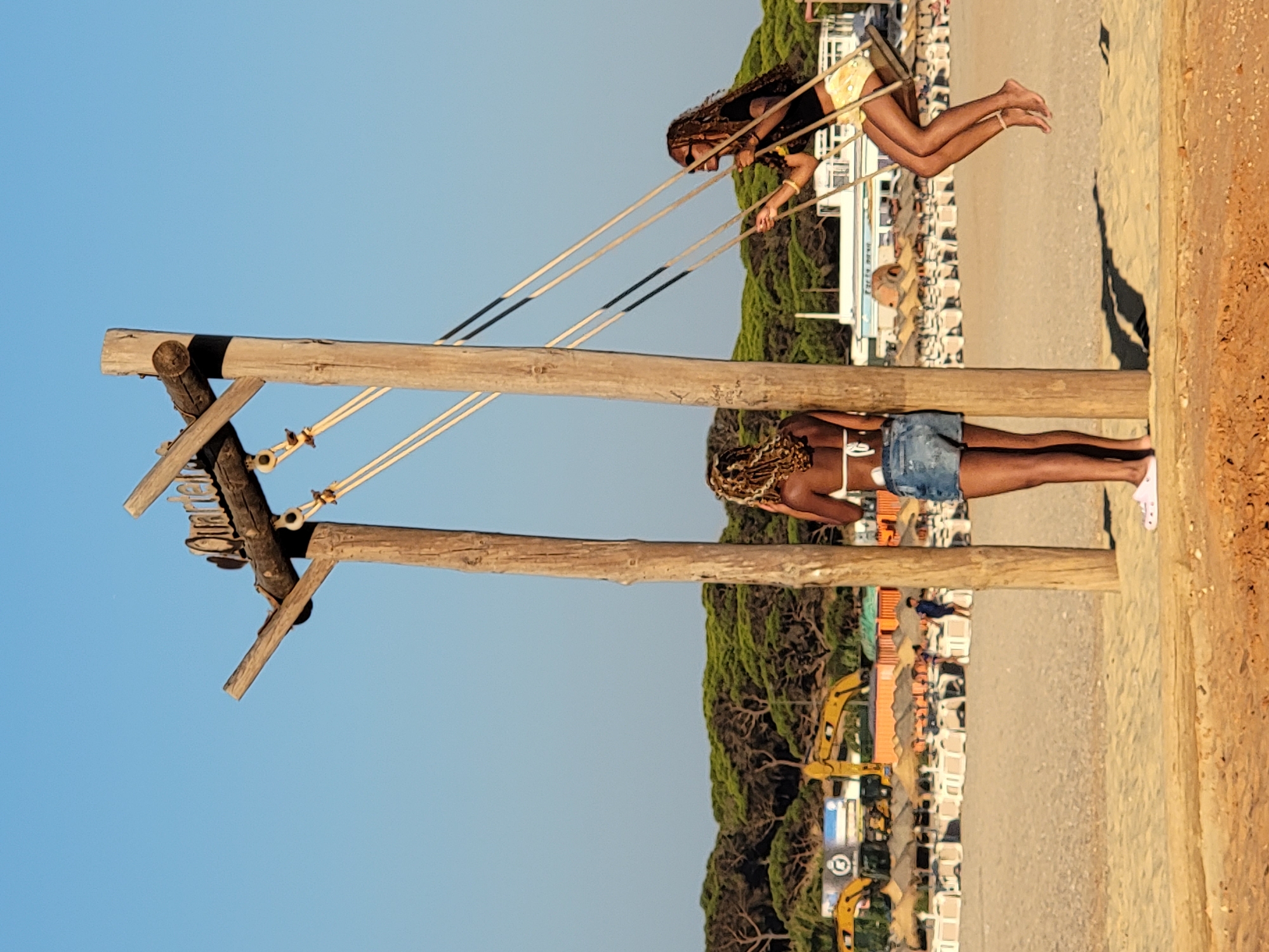 Two women are at the beach in Quarteira, Portugal. The woman on the right is swinging on a wooden swing attached to a wooden structure. She is wearing a black shirt and yellow shorts. The woman on the left is standing and facing away from the camera. She is wearing a white bikini top and denim shorts. In the background, there are beach umbrellas, lounge chairs, construction equipment, and trees. The sky is clear and blue.