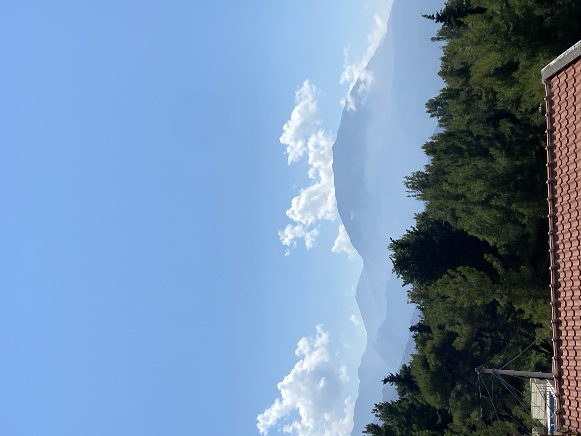 This is a landscape photograph taken in Šušanj, Montenegro, looking towards mountains on a clear day. The foreground features a red tiled roof and a line of dense green pine trees. Beyond the trees, a series of hazy blue mountains recede into the distance, with a larger, more prominent mountain peak partially obscured by clouds. The sky above is a vibrant blue, with scattered white cumulus clouds. There are no people or visible signs of activity in the image.