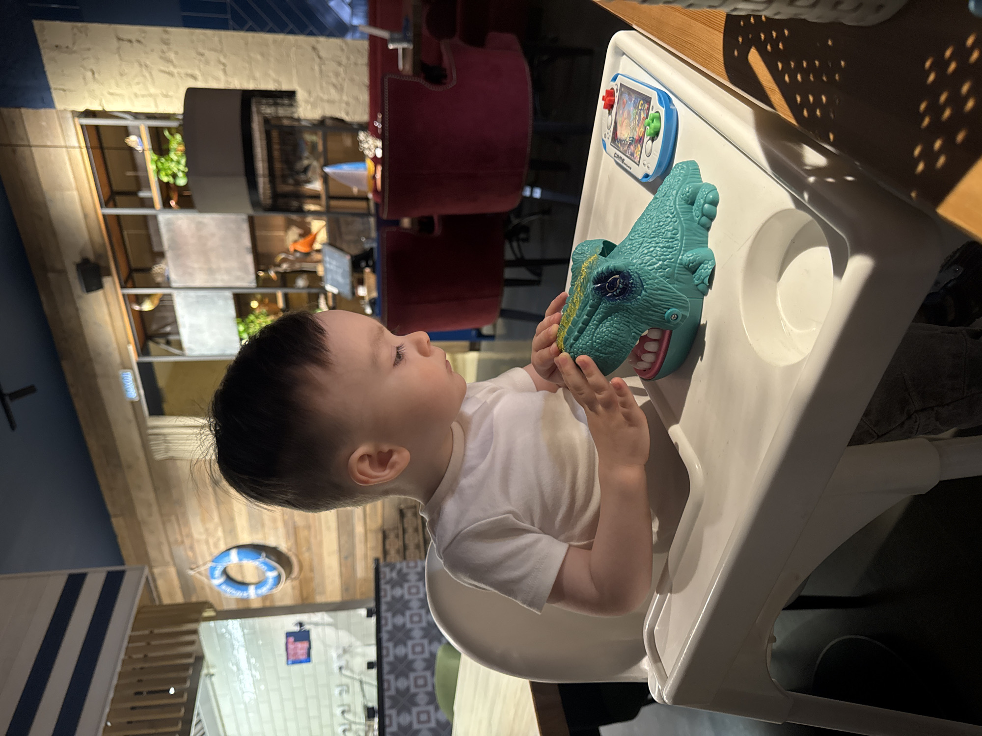 A young child sits in a white high chair, holding a blue dinosaur toy. To the left of the dinosaur toy is a white and blue handheld video game. The background features a restaurant interior, including maroon chairs, wooden paneling, shelves, and a lifebuoy decoration. The location is plausibly a restaurant in Izhevsk, Russia.