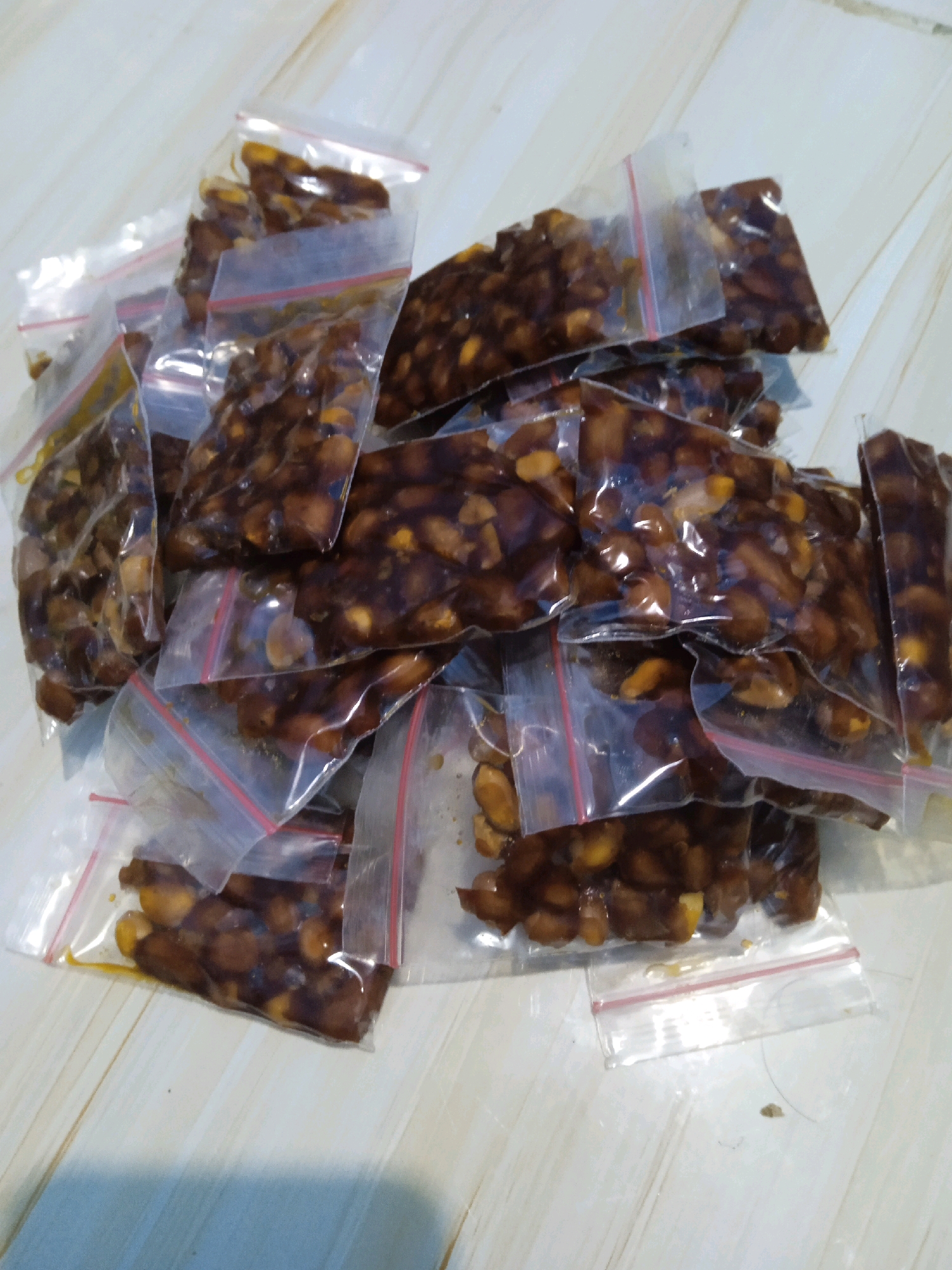 The image shows a pile of small, clear plastic bags containing what appears to be candied peanuts or a similar sweet snack. The bags are sealed with a red-lined zipper closure and are scattered across a light-colored, possibly wooden, surface with a wood grain pattern. The lighting is somewhat harsh, creating reflections on the plastic bags. The background is out of focus. There are no people visible in the image. The scene is an indoor setting, likely a home or a market stall, given the context of packaged food items. The location of Banjarmasin, Indonesia, does not appear to be discernible from the visual elements present in the image itself.