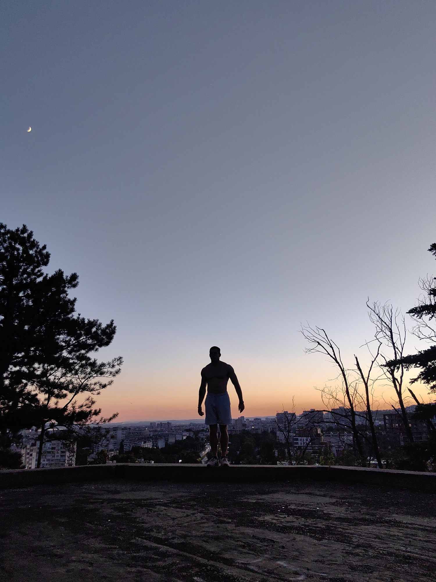 The image captures a silhouetted figure of a man standing on an elevated vantage point overlooking the city of Varna, Bulgaria. The time of day is dusk, with the sky displaying a gradient of colors from deep blue at the top to warm oranges and pinks near the horizon. A crescent moon is visible in the upper left portion of the sky.
The man is shirtless and wearing light-colored shorts, his back is to the camera, and he appears to be looking out at the cityscape. The foreground consists of a dark, textured surface, possibly asphalt or rough ground. To the left of the man, a dark silhouette of a pine tree stands tall. To the right, the bare branches of deciduous trees are silhouetted against the sky.
The city of Varna is spread out below, with buildings of varying heights visible. Some buildings appear to be apartment blocks, while others are less distinct. Streetlights or building lights are beginning to illuminate the city as the natural light fades. The perspective suggests the vantage point is on a hill overlooking the city, a common feature in Varna, such as the Asparuhov Bridge area or the Sea Garden hill. The overall atmosphere is serene and contemplative, emphasizing the vastness of the sky and the sprawling urban landscape.