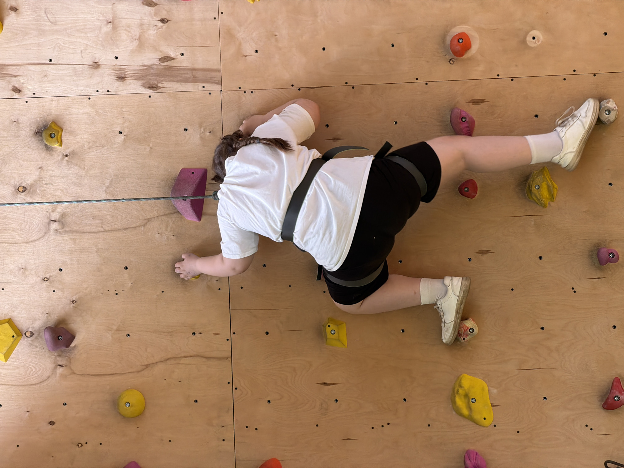 A young person wearing a white t-shirt, black shorts, white socks, and white sneakers is climbing a wooden indoor rock climbing wall. They are using a climbing harness and a rope. The wall has a variety of colored climbing holds. The location in Izhevsk, Russia, is not discernible from the image.