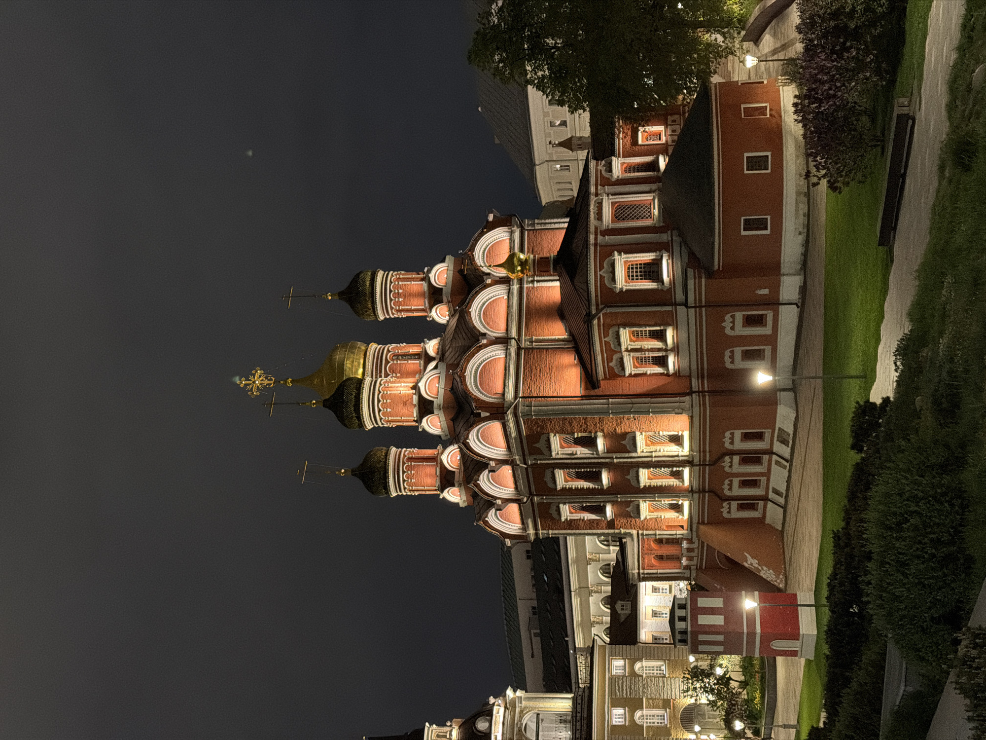 The image depicts a night scene in Moscow, Russia, featuring the Cathedral of the Icon of the Mother of God "Joy of All Who Sorrow" on Bolshaya Ordynka Street. The cathedral is illuminated, showcasing its distinctive onion domes with golden crosses. The surrounding area includes other buildings, trees, a paved walkway, and street lighting. The sky is dark, suggesting nighttime.