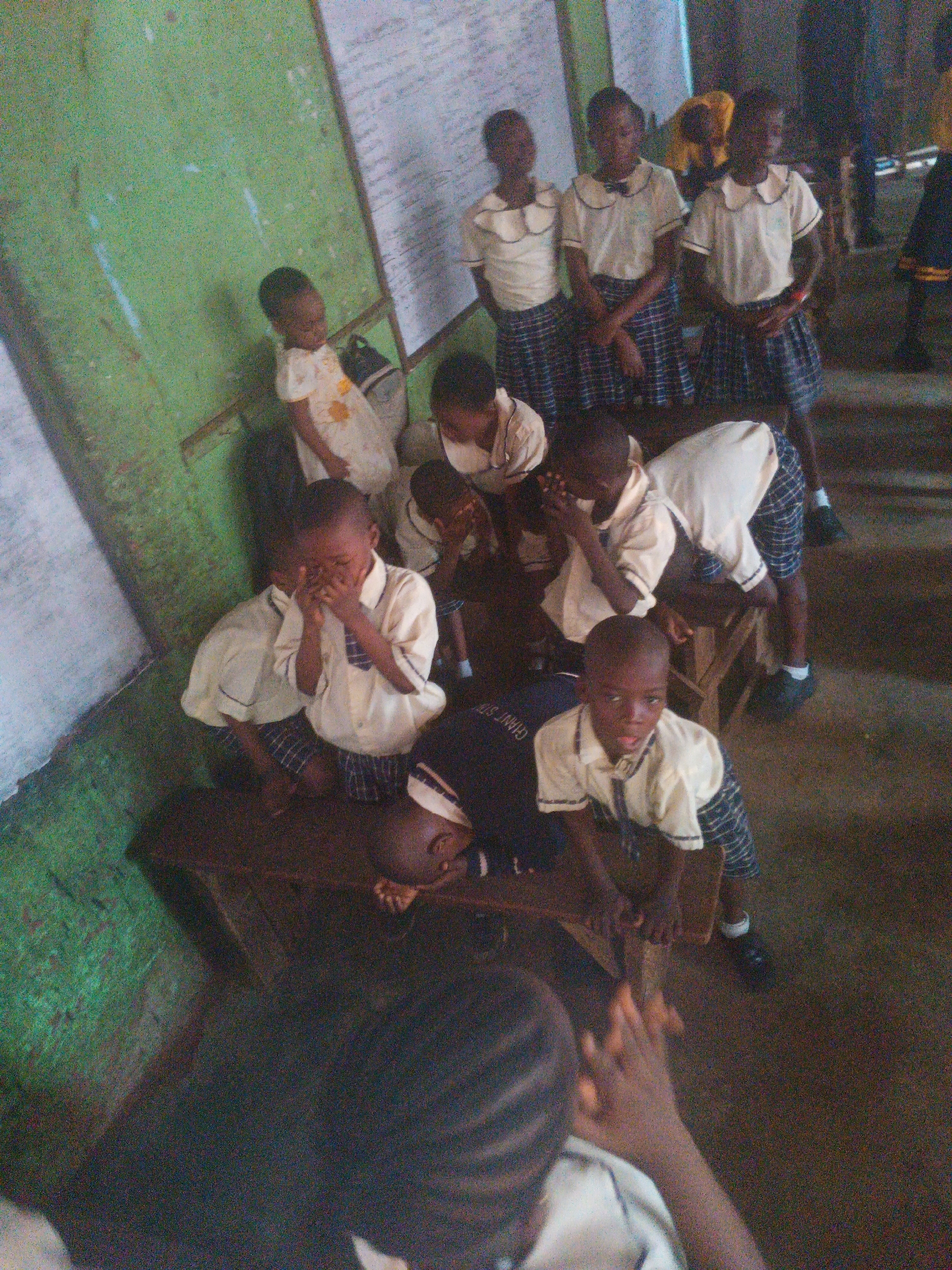 The image depicts a group of children, likely students, gathered in a classroom or similar indoor setting in Onitsha, Nigeria. The children are wearing school uniforms, consisting of white shirts and plaid skirts or shorts. Several children are seated on a wooden bench or table, while others are standing. Some children appear to be interacting with each other, while others are looking down or covering their faces, suggesting a moment of instruction, discipline, or perhaps distress. The environment appears somewhat worn, with a green painted wall and what looks like a whiteboard or bulletin board in the background. The overall impression is of a typical, albeit somewhat basic, educational setting for young children in Nigeria.