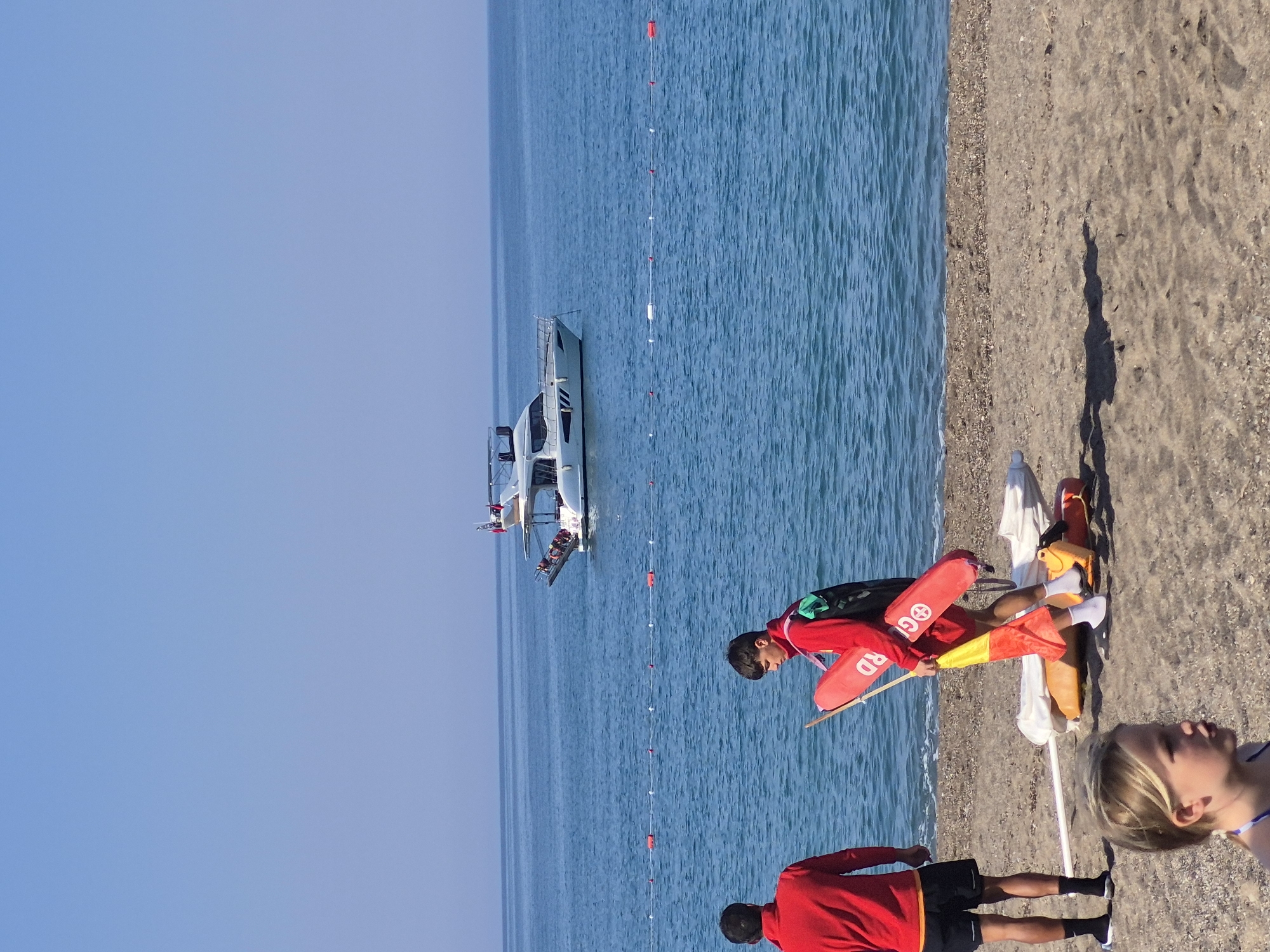 The image depicts a scene at a beach in Alanya, Turkey. The photo is rotated 90 degrees clockwise. The beach is sandy with a calm turquoise ocean visible. A line of red and white buoys stretches out into the water. A white boat filled with people is on the water. Two lifeguards in red shirts stand on the beach. One lifeguard is holding a red rescue float and a stick. A third person is lying on the beach. Beach umbrellas are also on the beach. The sky is clear and blue.
