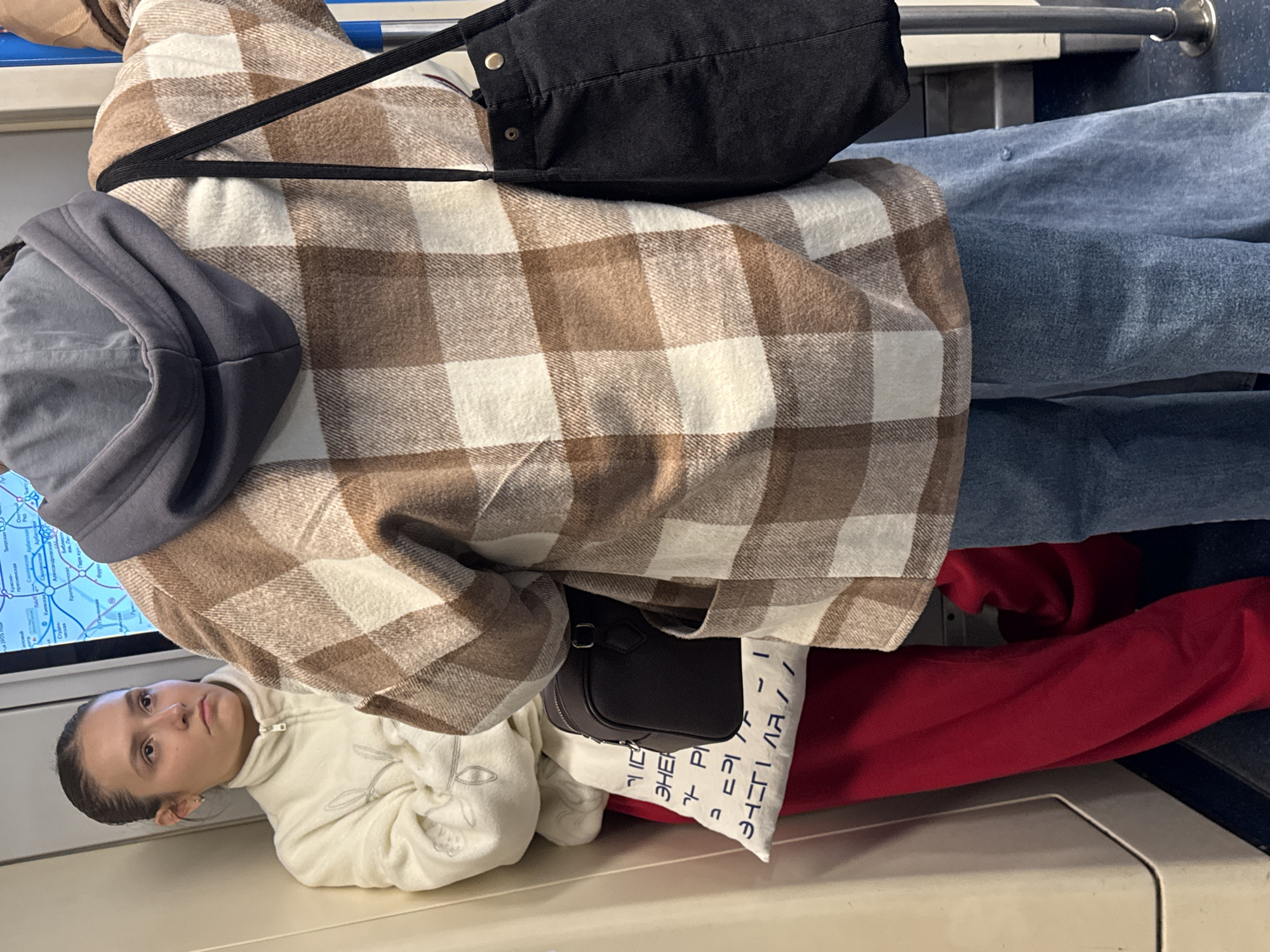 Two individuals are depicted inside a Moscow Metro train car, viewed from the rear and side. The person in the foreground wears a brown and white checkered jacket over a grey hoodie and light blue wide-leg jeans. They are carrying a black tote bag on their right shoulder. To the left, a young woman with her hair pulled back is leaning against the train car's wall. She is wearing a cream-colored fleece jacket with a zipper and red pants. She has a small dark brown handbag slung over her left shoulder. A white cloth bag with blue lettering is visible near her, tucked between her and the red pants. The train car interior is utilitarian, with visible metal poles and a map displayed on the wall behind the individuals. The lighting suggests an indoor, likely underground, environment.