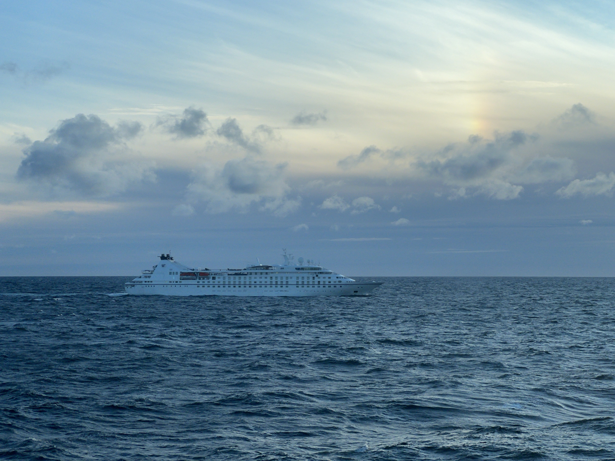 A large white cruise ship is sailing on a choppy, dark blue ocean. The ship is positioned in the lower right quadrant of the image and is oriented towards the left. The sky above is a gradient of light blue, with scattered clouds ranging from dark gray to white. A faint, colorful arc, possibly a sun dog or a weak rainbow, is visible in the sky above and slightly to the right of the ship. The overall lighting suggests it is either dawn or dusk. The location is described as Unknown, Poland.
