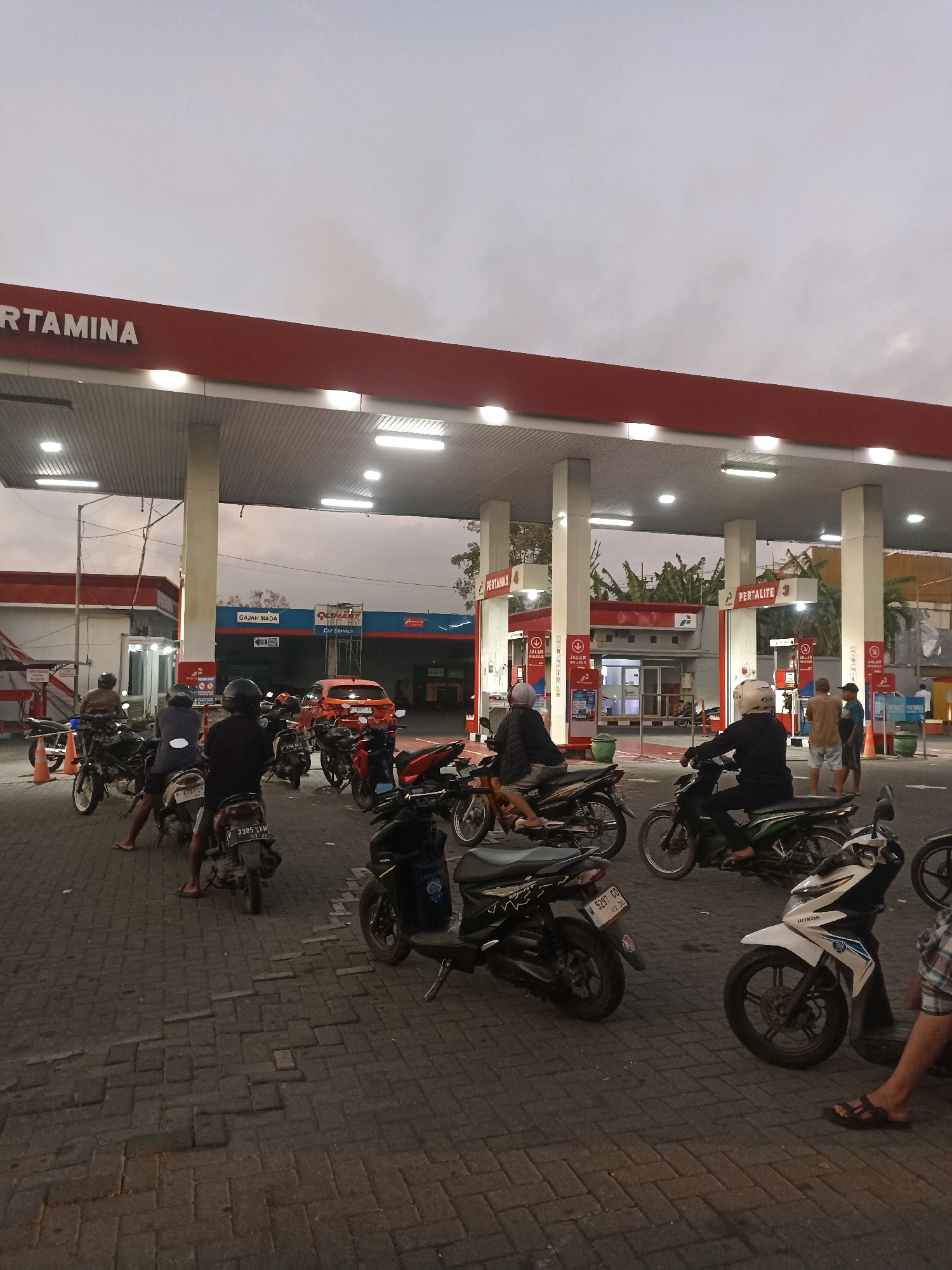 The image captures a scene at a Pertamina gas station in Banyuwangi, Indonesia, likely during dusk or early morning due to the ambient light. The gas station features a prominent red awning with white lettering reading "PERTAMINA." Underneath the awning, there are multiple white support columns and fluorescent lighting. Several people are present, predominantly on or around motorcycles, indicating a busy period at the station.
In the foreground, various motorcycles are parked or being operated. Some individuals are seated on their bikes, while others are standing or walking. The ground is paved with interlocking bricks. To the left, a building with red and white signage is visible, and an orange car is parked in the background. Further into the station, there are more pumps and signage, including "PERTALITE" and "PERTAMAX," with numbers 3 and 4 respectively. Two men in casual attire are standing near one of the pumps. The overall impression is of a typical, active scene at a gas station in an Indonesian city.