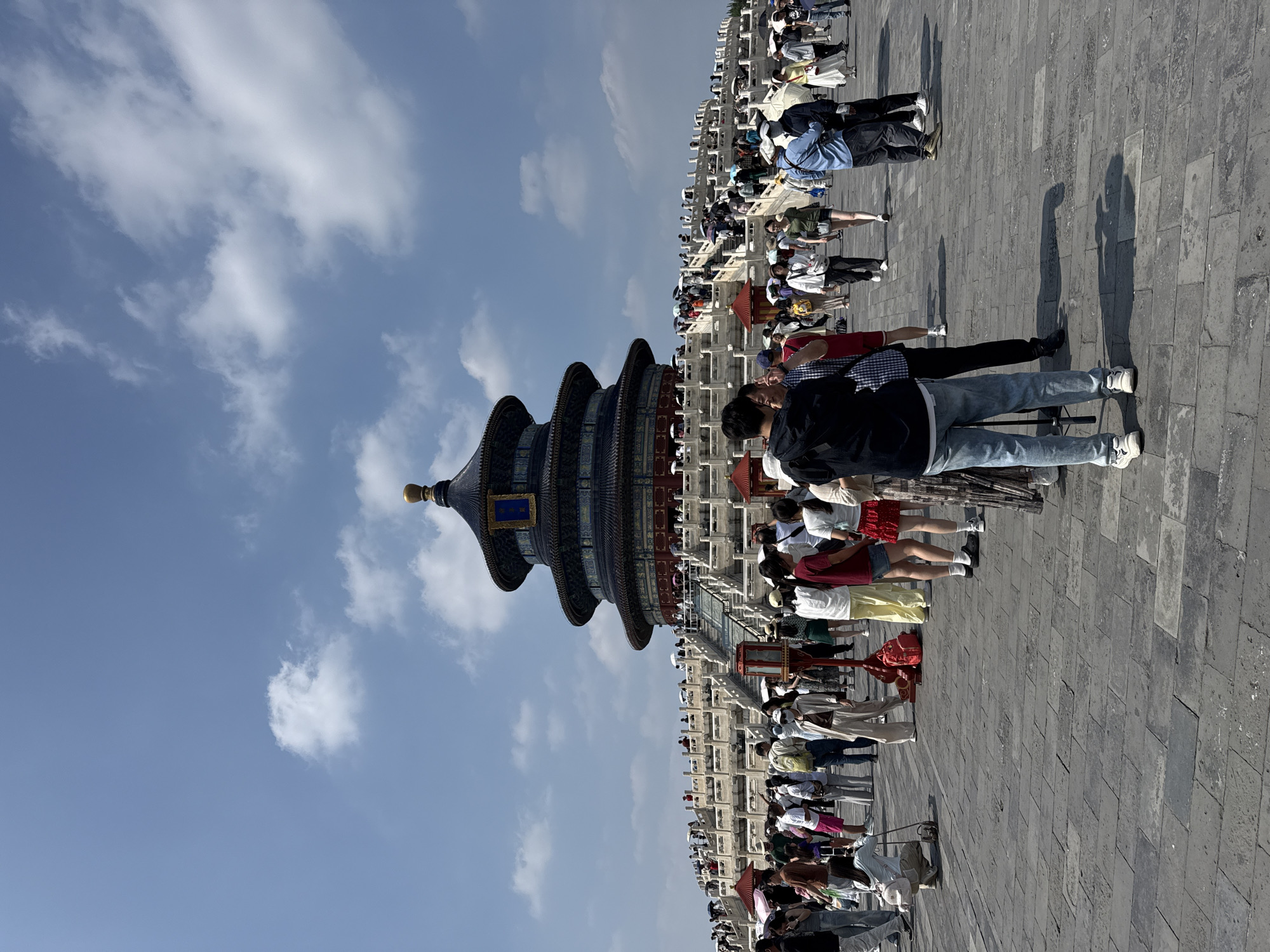 A vertical, eye-level outdoor shot captures the Hall of Prayer for Good Harvests at the Temple of Heaven in Dongcheng District, China, on a bright, cloudy day. The iconic, three-tiered, dark blue-roofed structure, adorned with gold accents, dominates the background. It is elevated on a multi-tiered, white marble terrace with ornate railings and a broad stone staircase leading up to it. Many visitors, some holding umbrellas, are visible on the terrace and the stairs. In the foreground, a large, grey paved plaza is filled with a diverse crowd of tourists. People are dressed in casual summer attire, including t-shirts, shorts, and jeans. Some are taking pictures, while others are walking or standing in groups. A few individuals are focused on their phones. The overall scene conveys a busy tourist destination.
