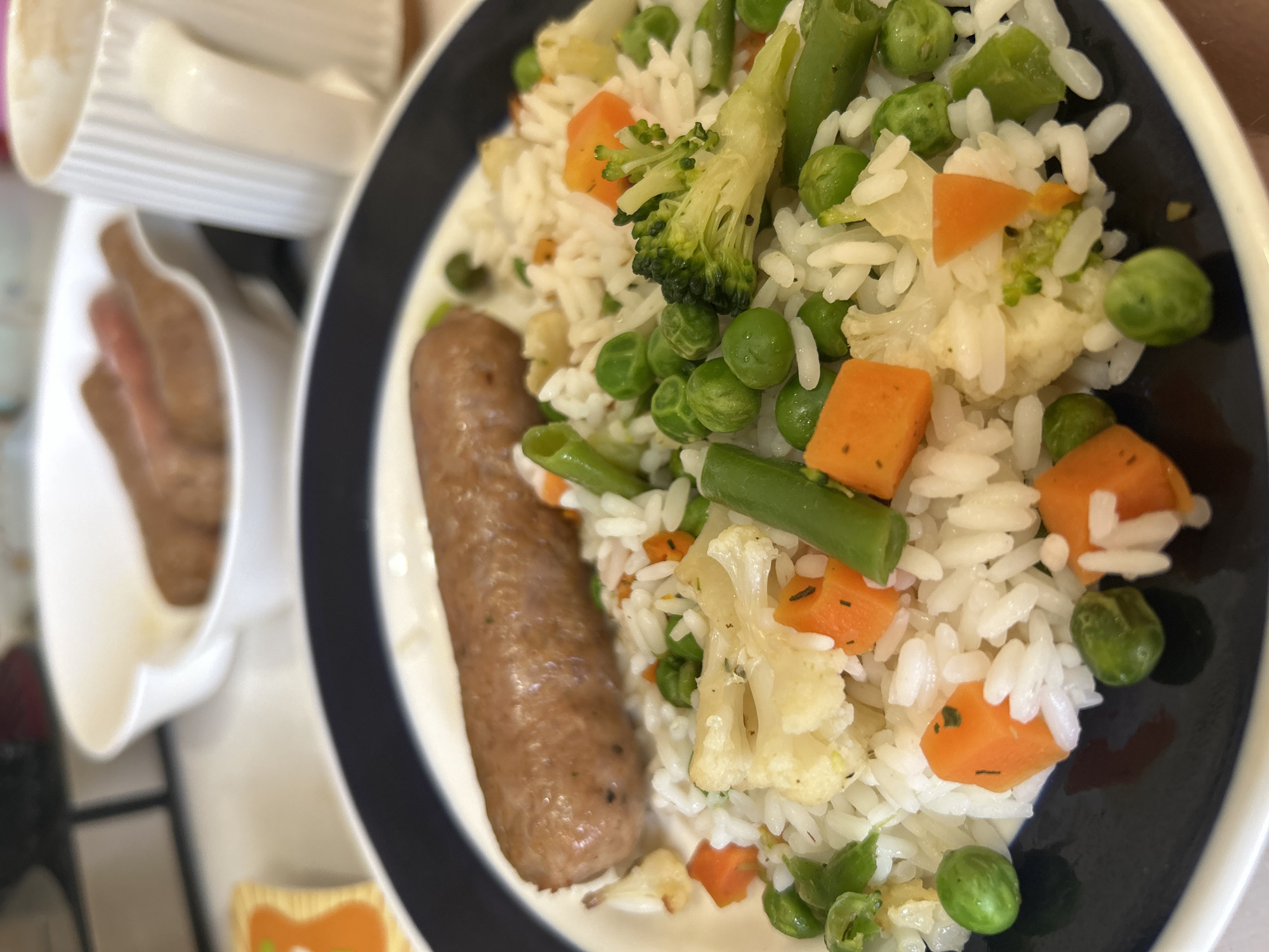 A plate of food consisting of white rice mixed with green peas, diced carrots, cauliflower florets, and green beans is presented alongside a sausage. A white ramekin dish containing sausage pieces is partially visible in the background, as are white cups and containers. The plate has a black rim, and the setting appears to be indoors. There is no indication of this being specifically in Chaikovsky, Russia.