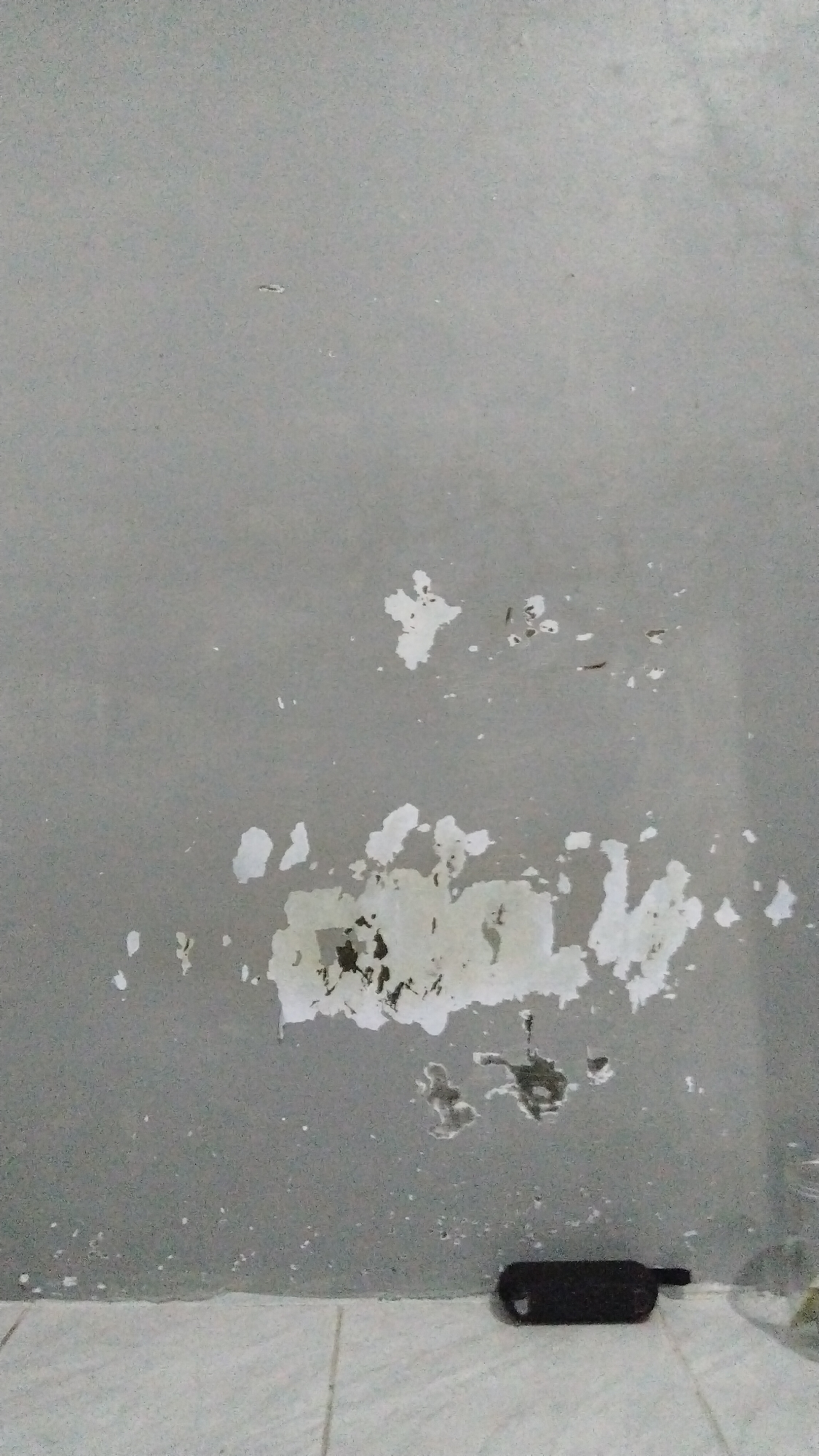 A gray wall in Banjarsari, Indonesia, exhibits patches of peeling paint, revealing darker plaster beneath. Some areas show small, scattered white specks and dark, irregular marks. A dark, cylindrical object, possibly a battery pack or a remote control, rests on the floor at the base of the wall. The floor is tiled with a light-colored, geometric pattern.