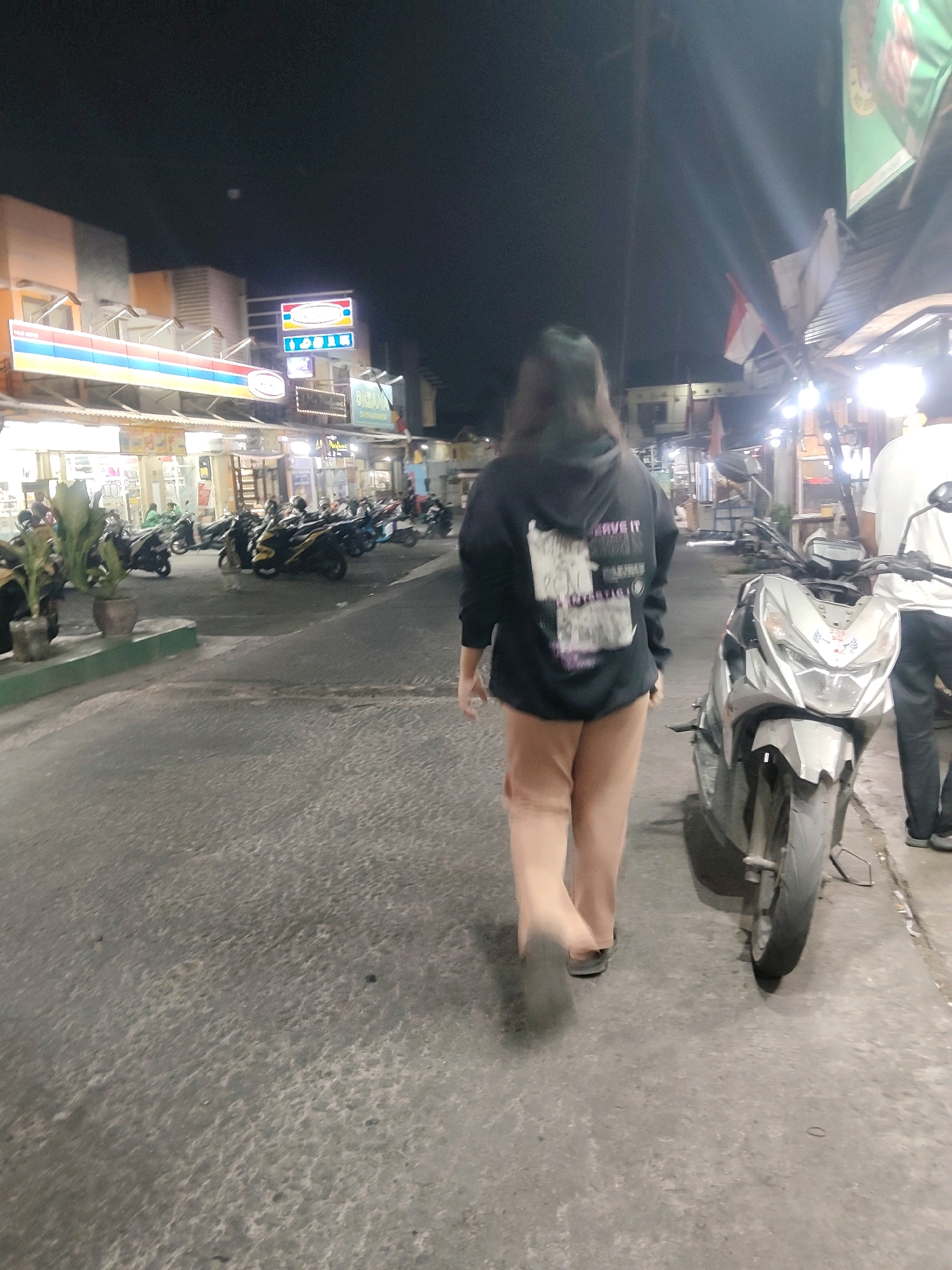 A rear view of a person wearing a black hoodie and light brown pants walks on a paved street at night in Mekarmukti, Indonesia. The person is walking away from the camera, towards several parked motorcycles. The street is lined with buildings with bright lights and signs, suggesting a commercial area or market. A motorcycle is parked to the right of the person, facing the camera. Another person wearing a white t-shirt and dark pants is visible in the background to the right, also walking away from the camera. The overall impression is a lively night scene in a city.