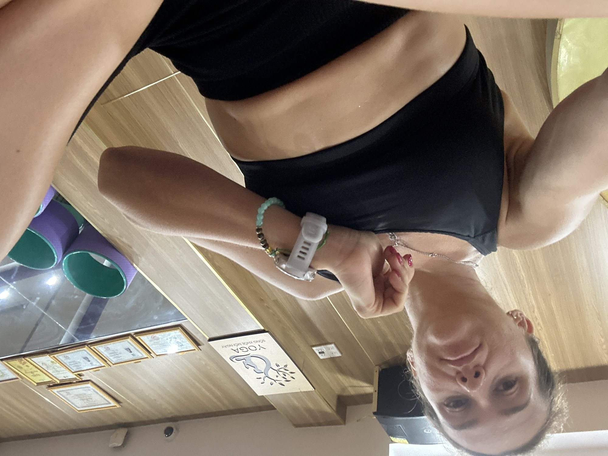A woman in athletic wear is positioned on the floor in what appears to be a yoga studio. She is looking up towards the camera with her head tilted. She has dark hair pulled back, and is wearing a black sports bra and black shorts. She is wearing a necklace, a bracelet with beads, and a fitness tracker on her left wrist. The background consists of a wooden wall with several framed certificates and a yoga studio sign. There are also yoga props visible, including colored foam rollers. The lighting suggests an indoor environment, possibly during the day. The image is taken from a low angle.