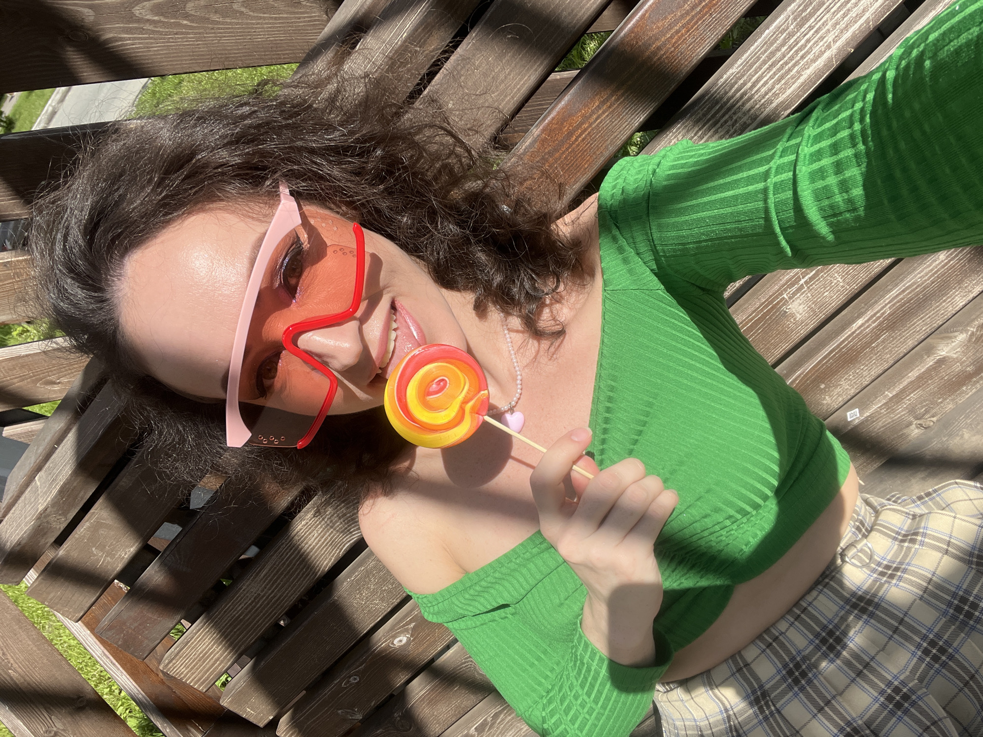 A woman is lying on a wooden bench outdoors, with grass visible in the background. She is wearing a green, off-the-shoulder top, a plaid skirt, and large pink-rimmed sunglasses with orange lenses. She is holding a colorful lollipop in her right hand and has her tongue sticking out slightly. The lighting suggests it is a sunny day, with shadows cast on the bench. No specific location in Kazan, Russia, can be identified from the image.