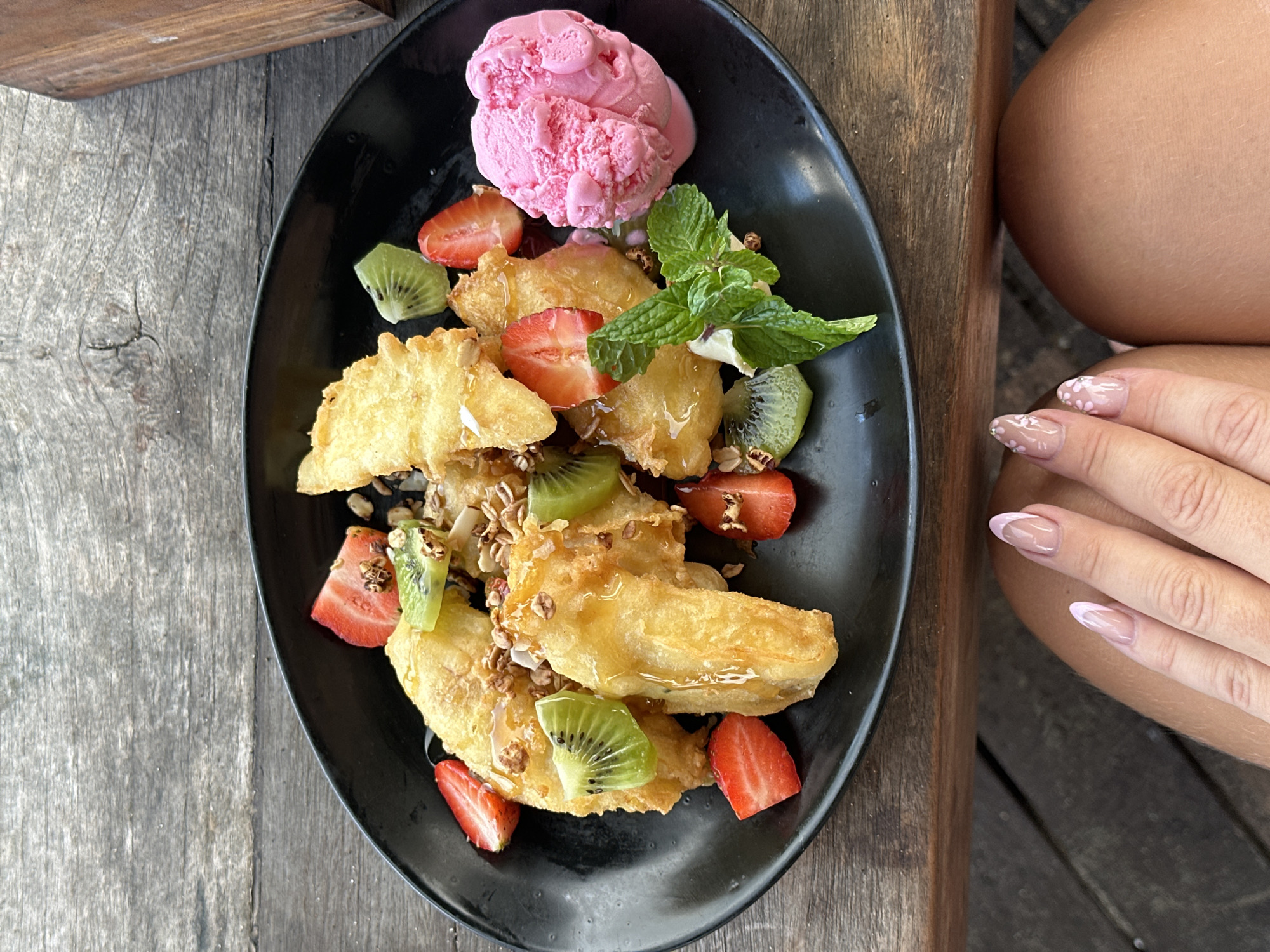 A close-up, overhead shot captures a black plate filled with golden fried banana fritters, garnished with fresh strawberries, kiwi slices, granola, mint leaves, and a scoop of bright pink ice cream. The plate rests on a weathered wooden table, likely outdoors given the natural lighting and the rustic wooden planks. In the foreground, a person's hand with light pink French manicured nails is visible, resting on their leg, suggesting a casual dining setting in the city of North Kuta, Indonesia, possibly at a cafe or restaurant with an outdoor seating area.