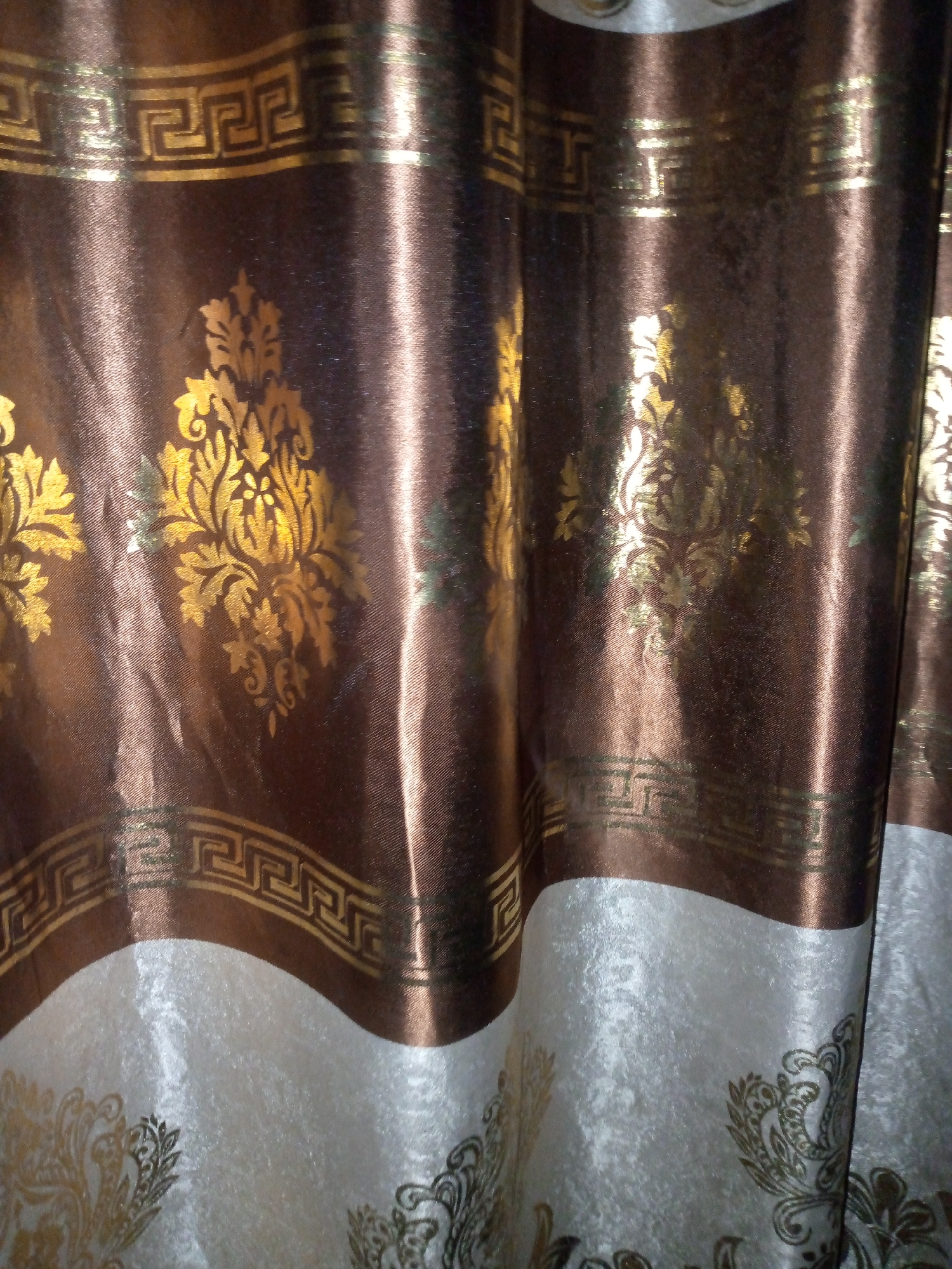 The image shows a close-up view of a decorative curtain with a metallic sheen. The curtain features a repeating Greek key pattern in gold along the top edge and a stylized floral motif, also in gold, on a rich brown background. The bottom portion of the curtain transitions to a lighter, textured fabric, possibly cream or off-white, with a complementary gold damask-like pattern. The lighting creates highlights and shadows on the textured fabric, emphasizing its softness and the metallic gleam of the gold accents. The folds of the curtain suggest it is hanging, likely in a room. There are no people or specific architectural details visible to pinpoint a location within the city of Khos, Uzbekistan.