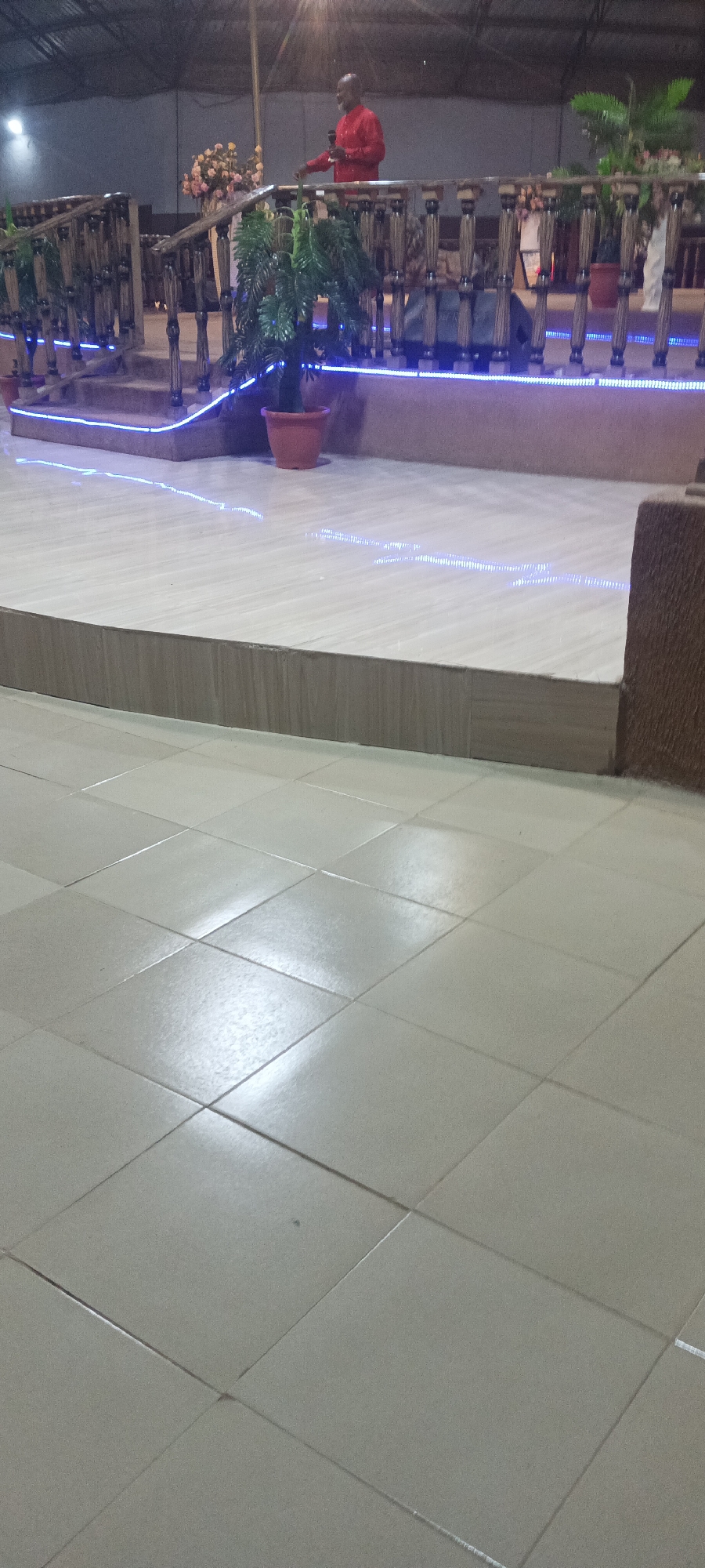 The image captures an indoor scene at what appears to be a church or public hall in Rukuba Road, Nigeria. The foreground shows a tiled floor with square-shaped tiles, while a raised platform is situated in the middle ground. This platform has a light-colored surface, trimmed with a darker horizontal panel. A railing surrounds the platform with decorative wooden posts. Blue LED lights outline the perimeter of the platform and the railing. Potted plants are visible at intervals along the railing.
In the background, an individual wearing a red shirt stands on the platform. The lighting is subdued, and the environment seems to be indoors under a roof structure. The scene is composed to emphasize the architecture of the platform and the floor space in front of it.