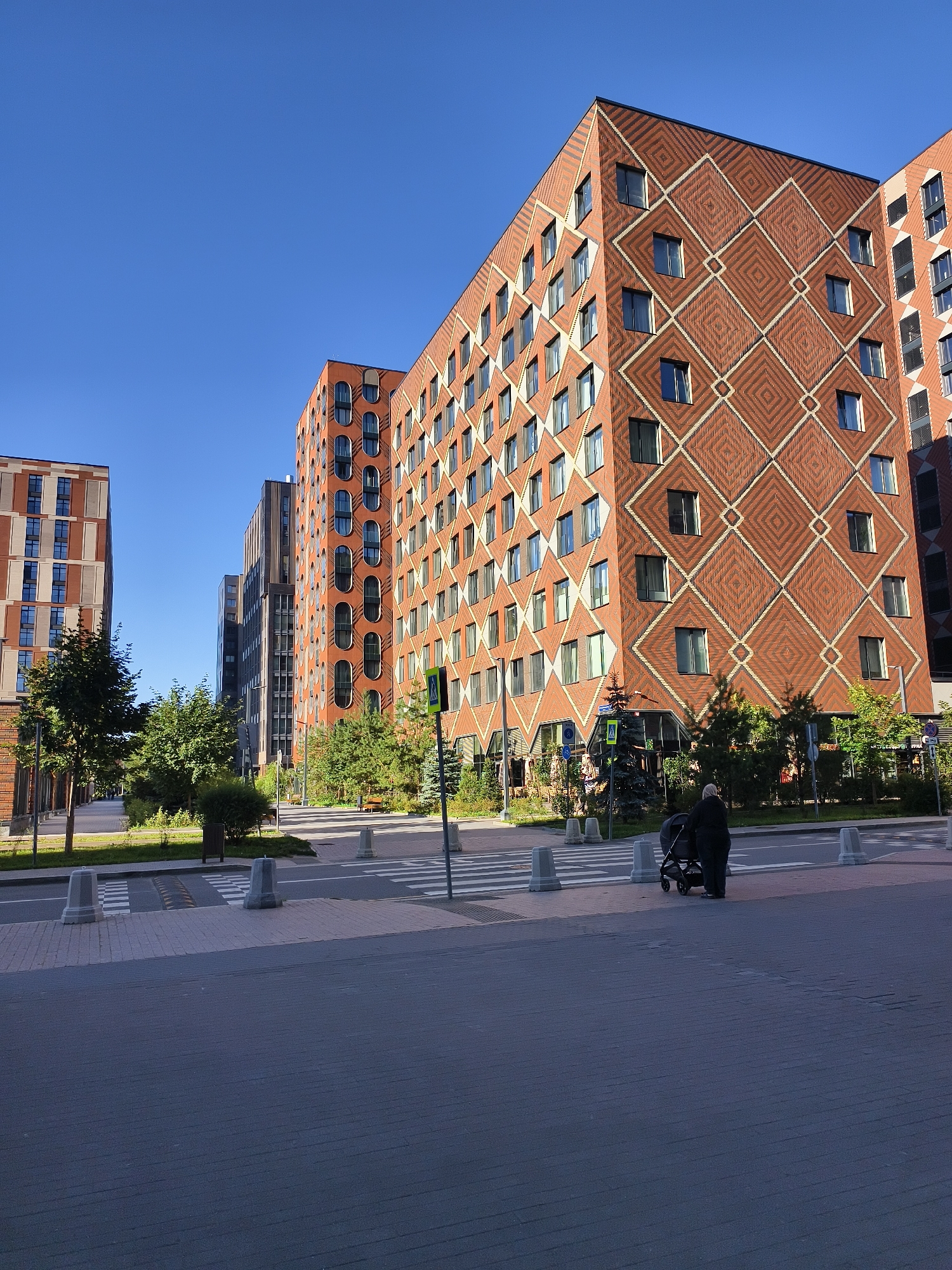 The media file shows a street scene in a modern urban environment, likely Moscow, Russia, under a clear blue sky, indicating a sunny day. The architecture features several tall residential buildings with distinct, patterned facades. One prominent building displays a striking geometric pattern of diamond shapes in reddish-brown brickwork with lighter accents. Another building to its left has a more linear pattern of red and beige brick. In the background, a darker, taller building with a glass facade is visible.
The foreground shows a pedestrian crossing with gray concrete bollards and a speed bump. A person, whose gender is not definitively clear but appears to be an adult, is pushing a dark-colored baby stroller across the street. They are wearing dark clothing and a head covering, partially obscuring their face. Lush green trees and bushes line the sidewalk and the areas between the buildings, suggesting a well-maintained urban landscape. The scene appears calm and everyday, with the focus on the residential buildings and the pedestrian activity. No visible text is present in the media file. The time of day is likely morning or afternoon, given the clear sunlight and the shadows cast.