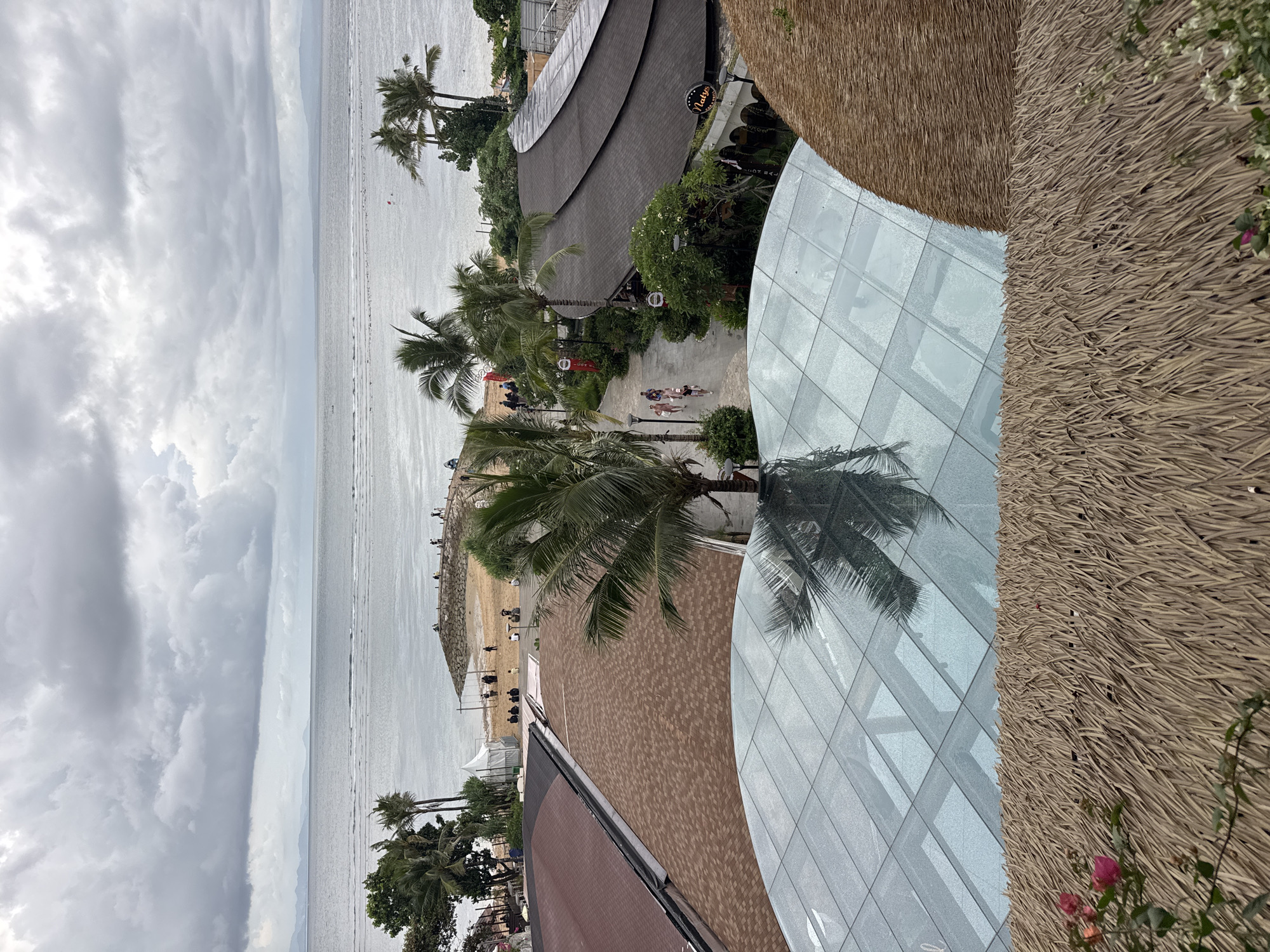 An elevated view captures a tropical beachfront resort area under an overcast sky. The image depicts a dynamic blend of natural coastal beauty and modern resort architecture, consistent with a setting in Denpasar, Indonesia.
On the left side of the frame, a wide expanse of cloudy grey sky transitions into a calm, silvery ocean with subtle ripples. A strip of sandy beach separates the ocean from the developed area. In the far distance, a faint coastline is visible beneath the heavy cloud cover.
The middle and right portions of the image feature the resort grounds. Numerous lush palm trees are scattered throughout, with some reflecting off a large, curved glass-paneled building in the foreground on the right. Adjacent to this glass structure is a section of a rough, brown thatched wall or roof, adding a traditional aesthetic. Further back, a paved pathway winds through the resort, where several small groups of people are seen leisurely walking. Another prominent, curved building with a dark, shingled roof stands beside the pathway, featuring a circular sign with the text "Uluwatu" clearly visible. On the beach, a few small tents and individuals are present, and a stone jetty or breakwater extends into the ocean, with more people gathered on it.
The time of day is daytime, with soft, diffused lighting characteristic of an overcast sky. The overall atmosphere suggests a relaxed, leisurely environment at a tropical destination.