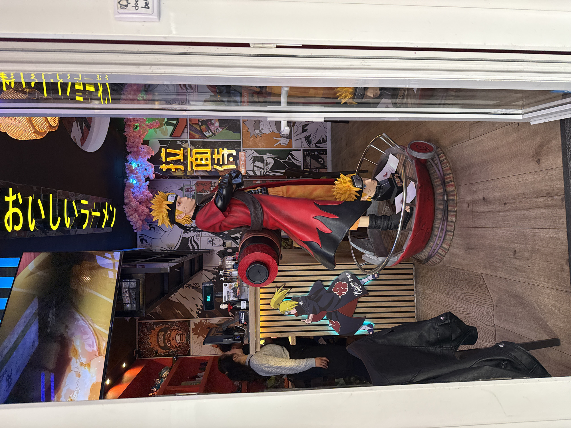A window display in Budapest, Hungary features anime-themed elements. A Naruto figure is centrally positioned on a red stand. Behind the figure are manga-style posters. A neon sign with Japanese text is visible. Another character sticker is positioned at the base of the window display, at a lower level. A person wearing a light colored sweater and black pants stands inside the restaurant, looking into the window. A black leather jacket lays beside them. The floor is made of wood. A small sign with text is affixed to the upper left of the window.