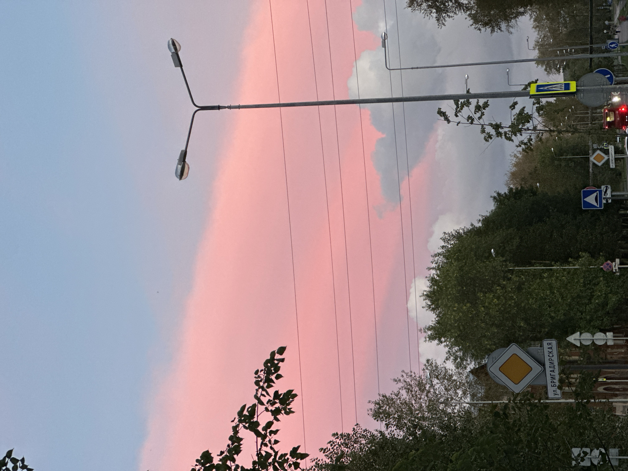 The image depicts a street scene in Perm, Russia, under a pink and blue sky with scattered clouds, likely at dusk or dawn. A prominent lamppost with two lights stands near the center, with other streetlights and power lines visible. Lush green trees line the street, and several traffic signs are present, including a yield sign with Cyrillic text that reads "ул. БРИГАДИРСКАЯ" (Brigadirskaya Street). The street itself is paved, and a red vehicle with illuminated taillights is visible in the distance. The overall atmosphere is tranquil, with a blend of urban elements and natural scenery.