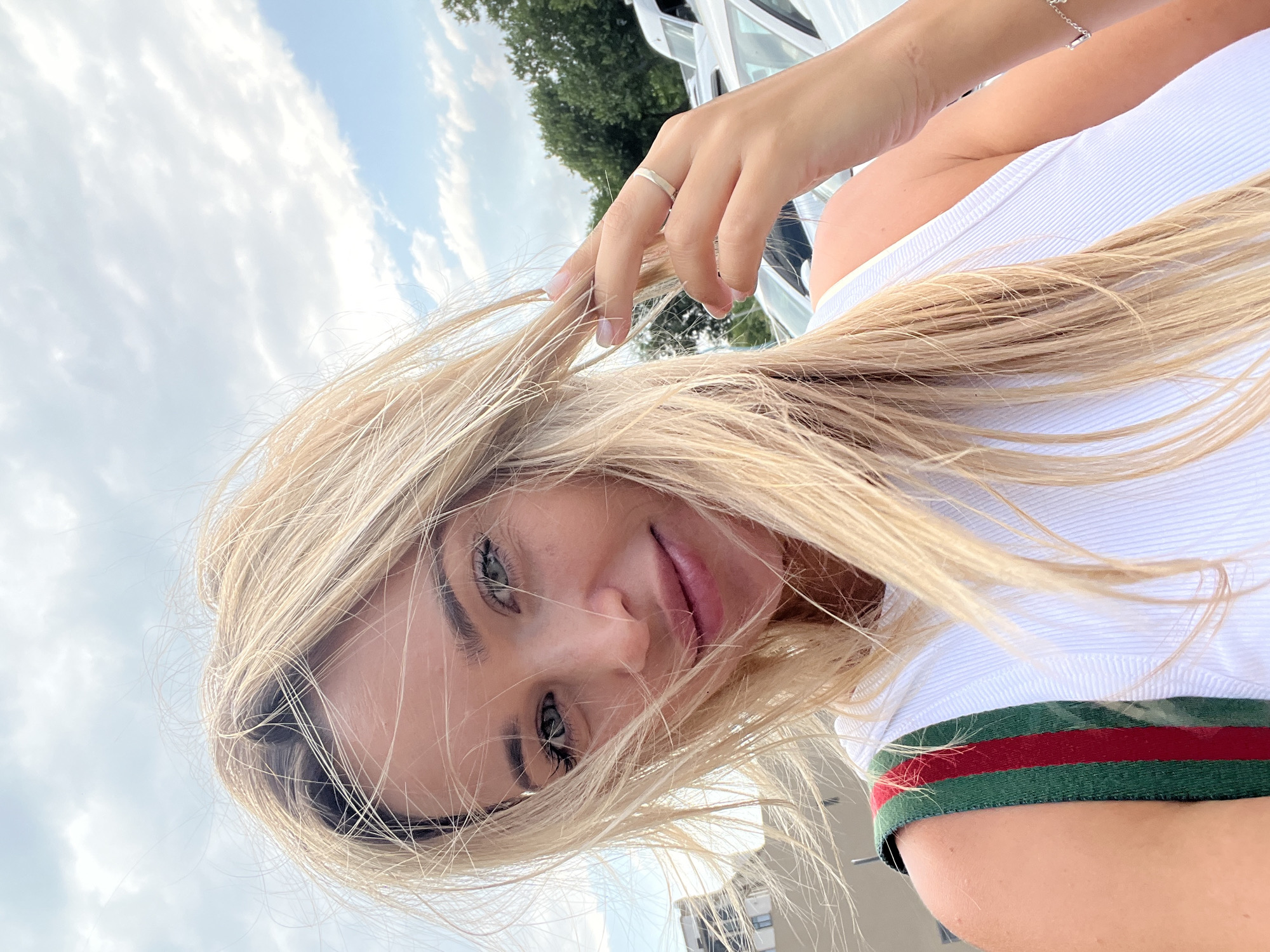 An outdoor, medium shot, selfie-style photograph shows a blonde woman with wind-blown hair. She is wearing a white, ribbed tank top with red and green striped straps. Her left hand is touching her hair, and she is wearing a ring on her right ring finger. In the background, there are parked white vans, trees, and a cloudy sky. The location is not specified as Fontanka, Ukraine.