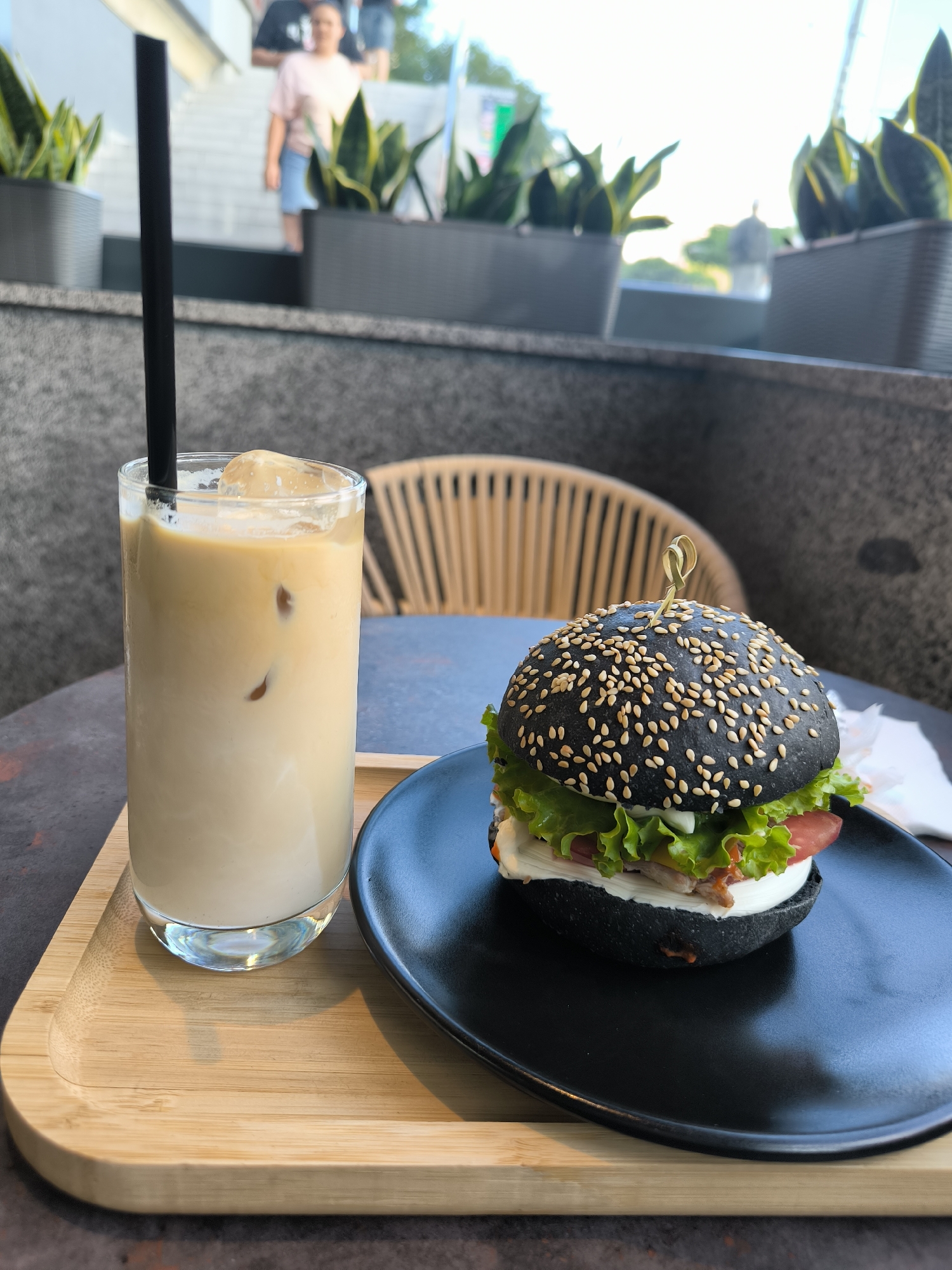 An eye-level, close-up shot captures a black sesame seed bun hamburger and a tall glass of iced coffee on a table. The hamburger, skewered with a wooden pick, sits on a black plate. Its fillings, including lettuce, tomato, and a white sauce, are visible. The iced coffee, served in a tall glass with a black straw and ice cubes, is placed on a wooden tray next to the hamburger. In the background, blurred figures of people are visible near a staircase and potted plants, suggesting an outdoor seating area of a cafe or restaurant. The lighting suggests it is daytime.