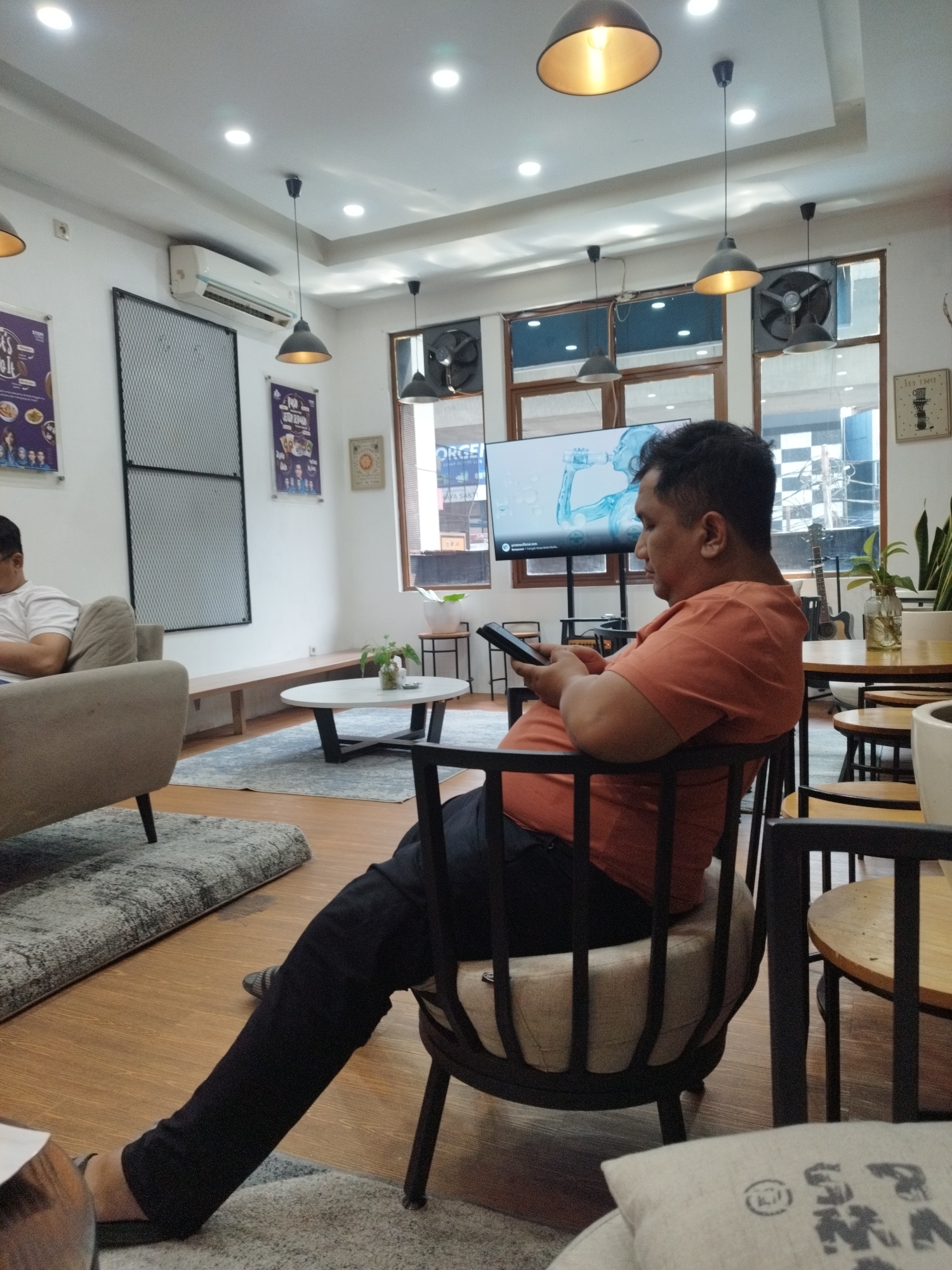 The image shows the interior of a cafe in South Jakarta, Indonesia. A man in an orange t-shirt and black pants is seated on a chair, looking at his phone. He is in the foreground, with a sofa and a rug to his left. In the background, there is a television screen displaying an advertisement, a coffee table with a plant, and several windows. The cafe appears to be modern and well-lit, with an air conditioner mounted on the wall. The overall atmosphere is relaxed and casual.