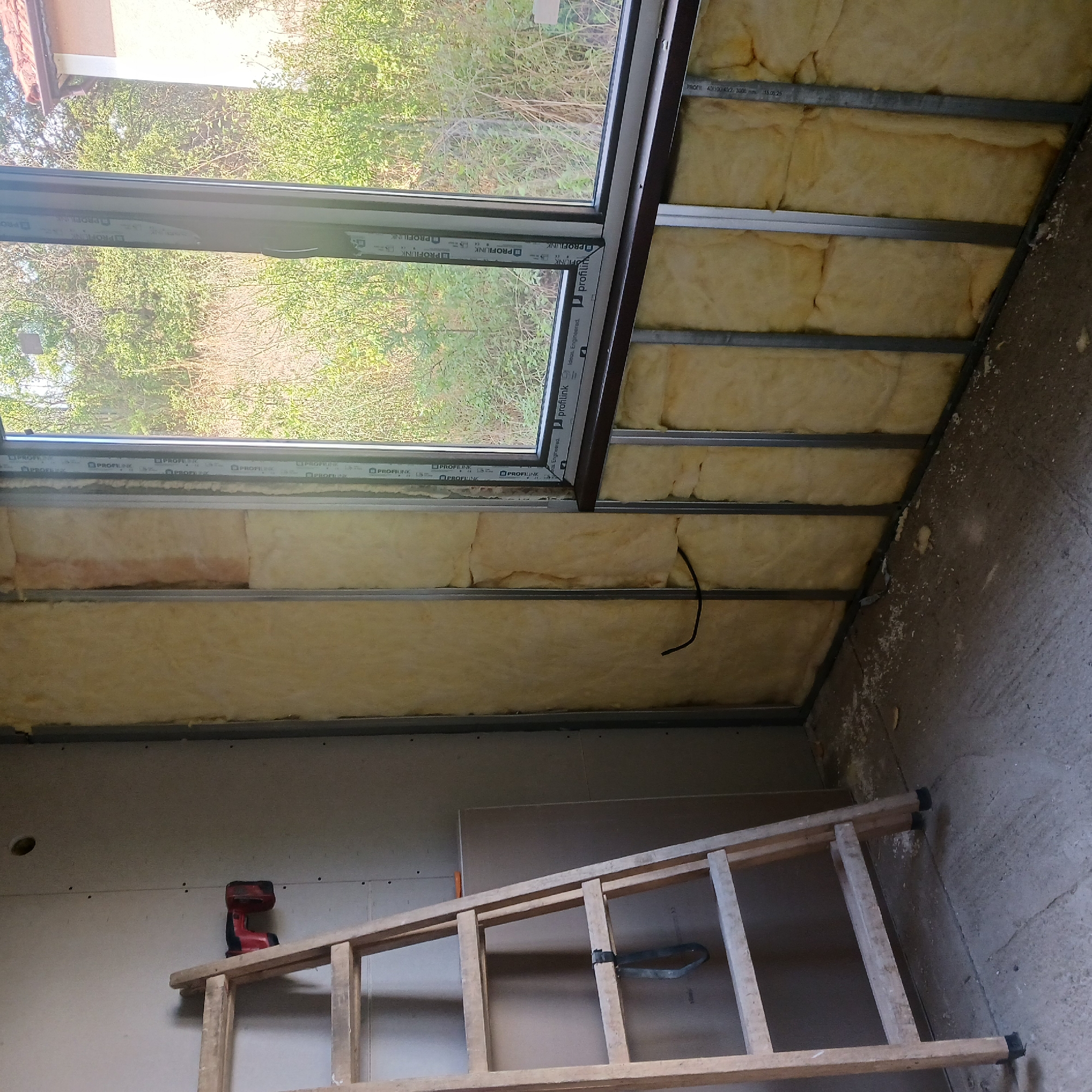 An interior view shows a room under construction in Ostra Mogila, Bulgaria. A wooden ladder leans against a wall covered in drywall, where a red power drill is attached. The adjacent wall is framed with metal studs and filled with yellow insulation material, revealing a window overlooking a wooded outdoor area. The floor is concrete and shows debris. The overall scene depicts a residential construction site.