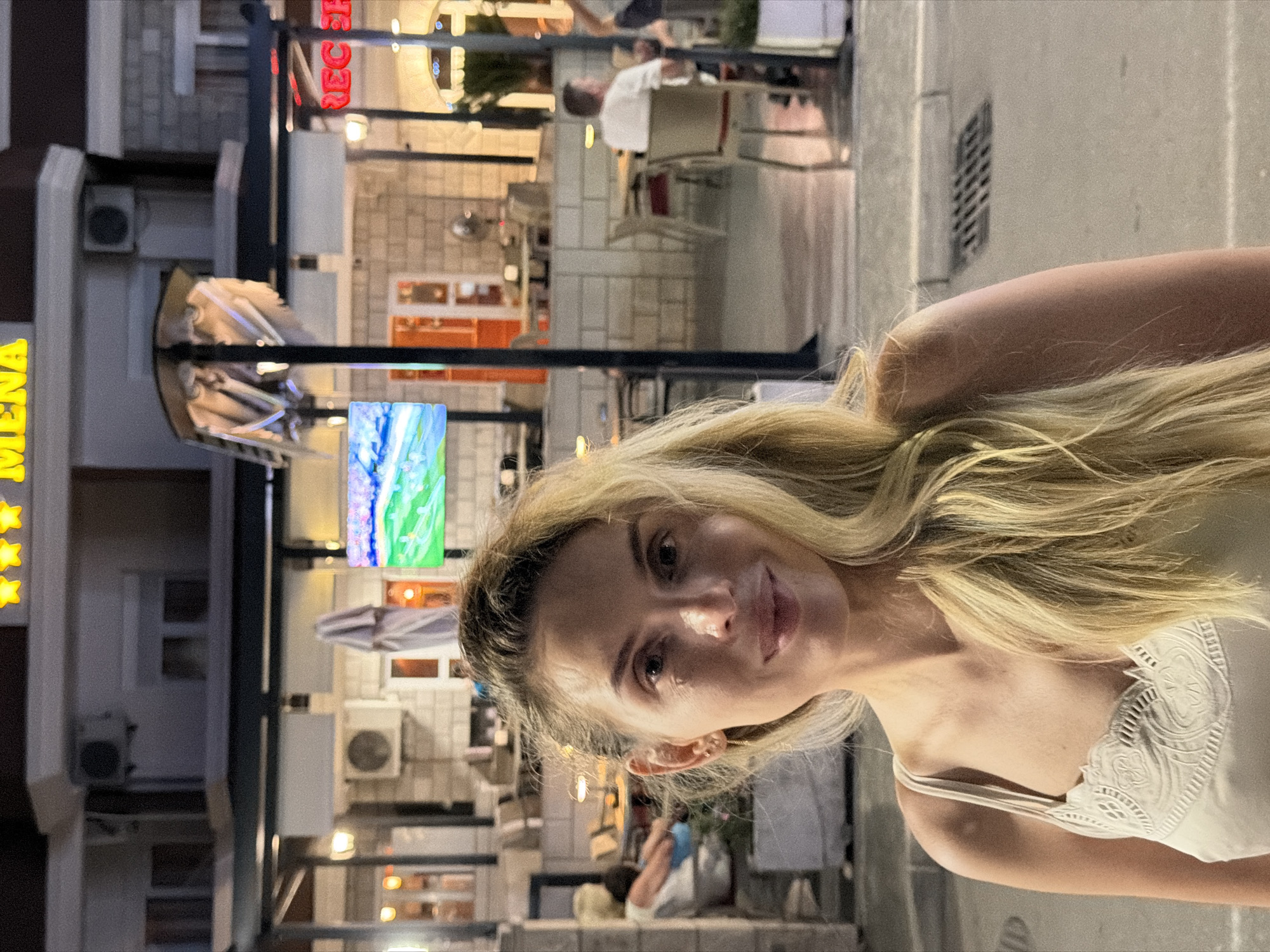 In Budva, Montenegro, a blonde woman with light-colored eyes and fair skin is pictured in front of a brightly lit establishment. She is wearing a light-colored tank top with intricate white lace detailing on the bust. Her hair is long and blonde, with some strands falling across her face. The background reveals an outdoor seating area of a restaurant or cafe. Tables and chairs are visible, with patrons seated and interacting. The establishment's facade features large windows and a prominent sign displaying the word "MENA" in yellow capital letters at the top, and "REC EF" in red capital letters on the right side. Other details include an umbrella in the foreground, an air conditioning unit mounted on the building, and a drain grate on the pavement. The lighting suggests it is evening.