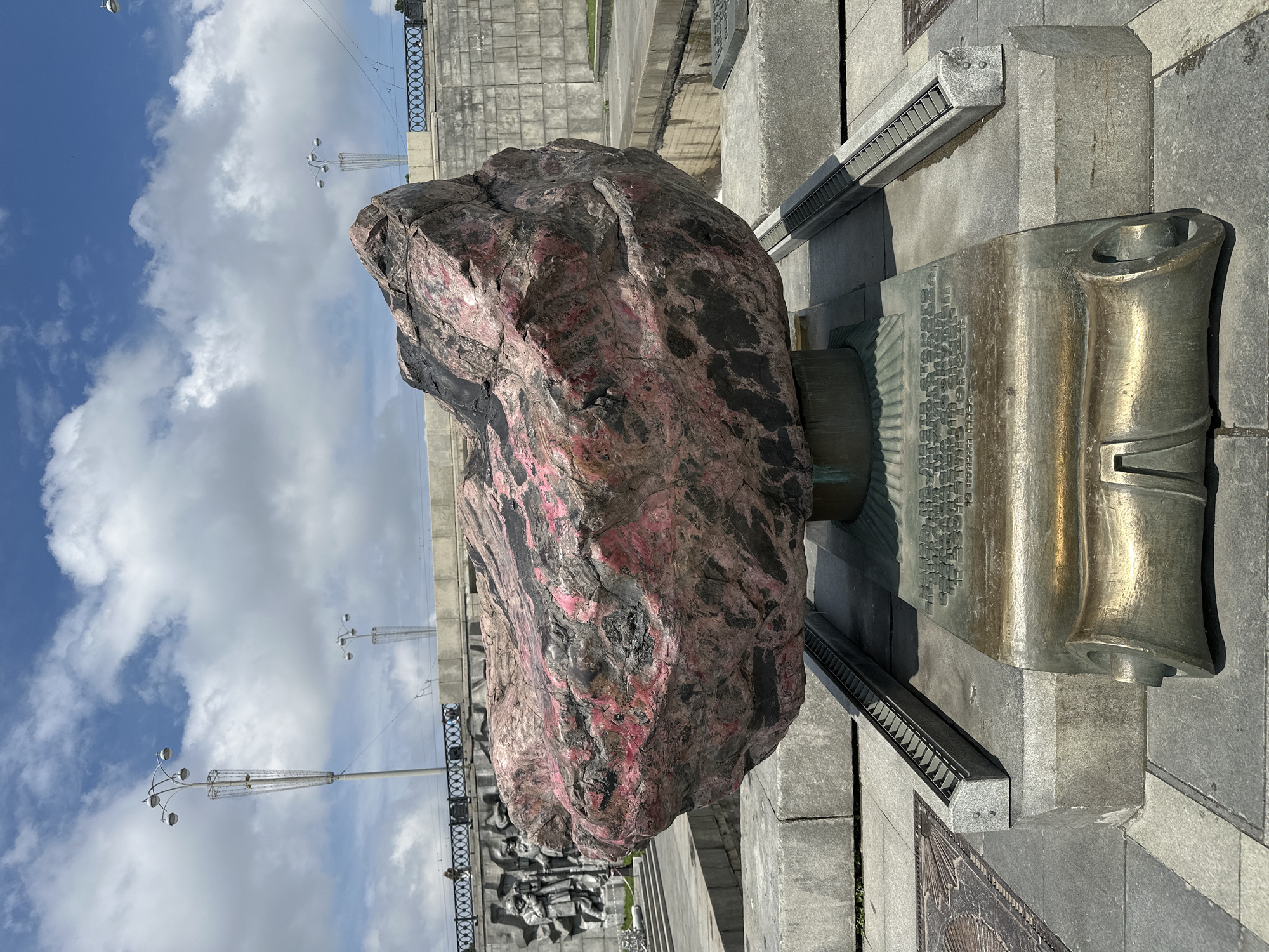 A large, irregularly shaped boulder, predominantly gray and pink with black veining, rests on a cylindrical pedestal. The boulder is positioned in the foreground of the image. Below the boulder, a bronze plaque with engraved text is mounted on a stone base. The stone base is part of a larger monument, which includes recessed channels on either side, filled with what appears to be a metal grate. In the background, a wall with bas-relief sculptures of figures and a decorative metal railing are visible. Two tall lampposts with multiple lights stand on either side of the railing. The scene is set outdoors under a cloudy blue sky. This monument is the "Stone of Friendship" located on the embankment of the Iset River in Yekaterinburg, Russia, commemorating the friendship between the city and the Czech Republic.