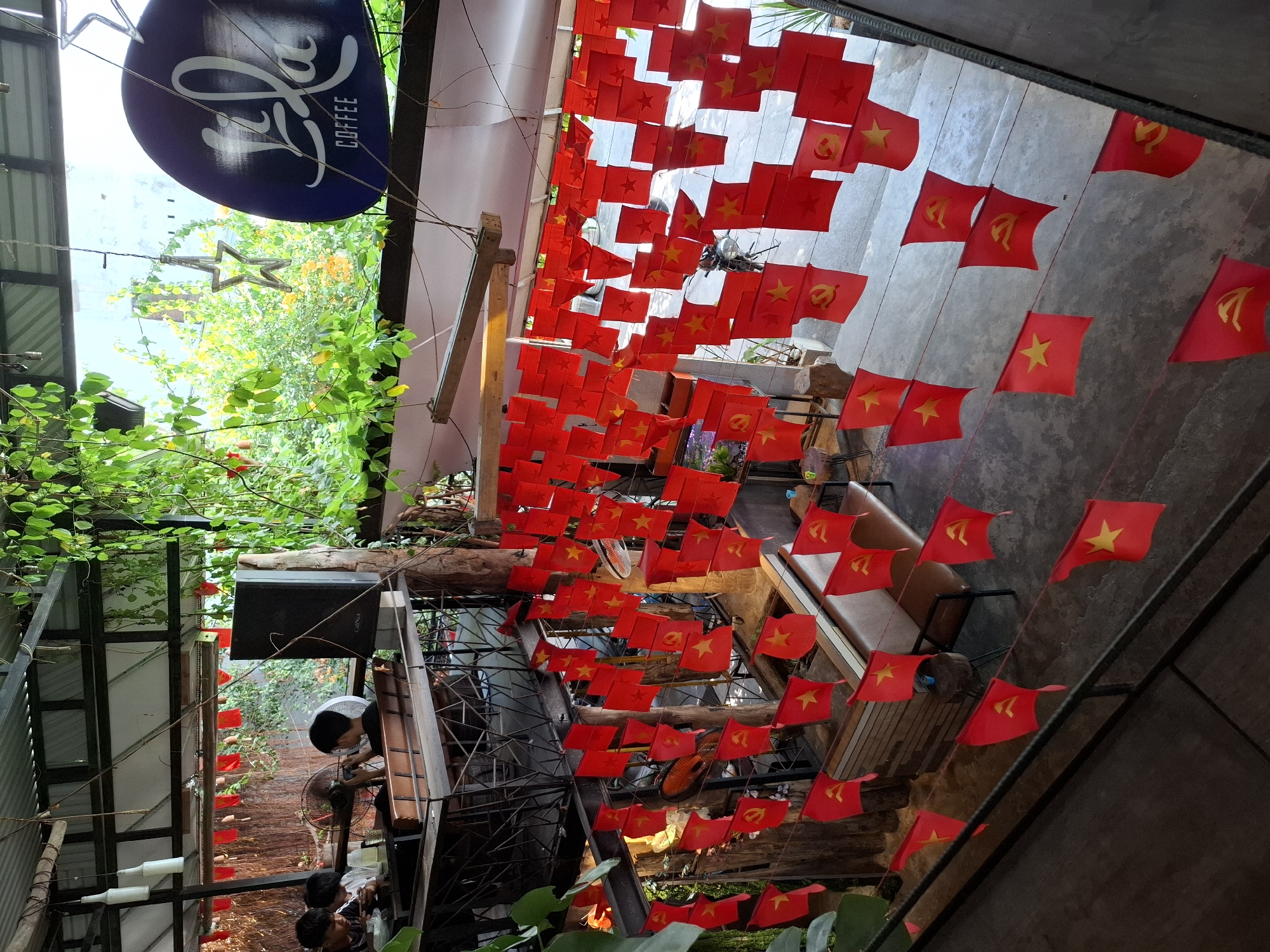 This image shows the interior of a coffee shop in Nha Trang, Vietnam. The shop is decorated with many small red flags, some featuring a yellow star and others featuring a hammer and sickle. There are plants hanging from the ceiling and walls, creating a lush, green atmosphere. Several people are visible, sitting at tables and looking at their phones. The furniture is mostly wooden and metal. The lighting is somewhat dim, but natural light is coming in from the open ceiling area. The overall ambiance appears casual and relaxed.