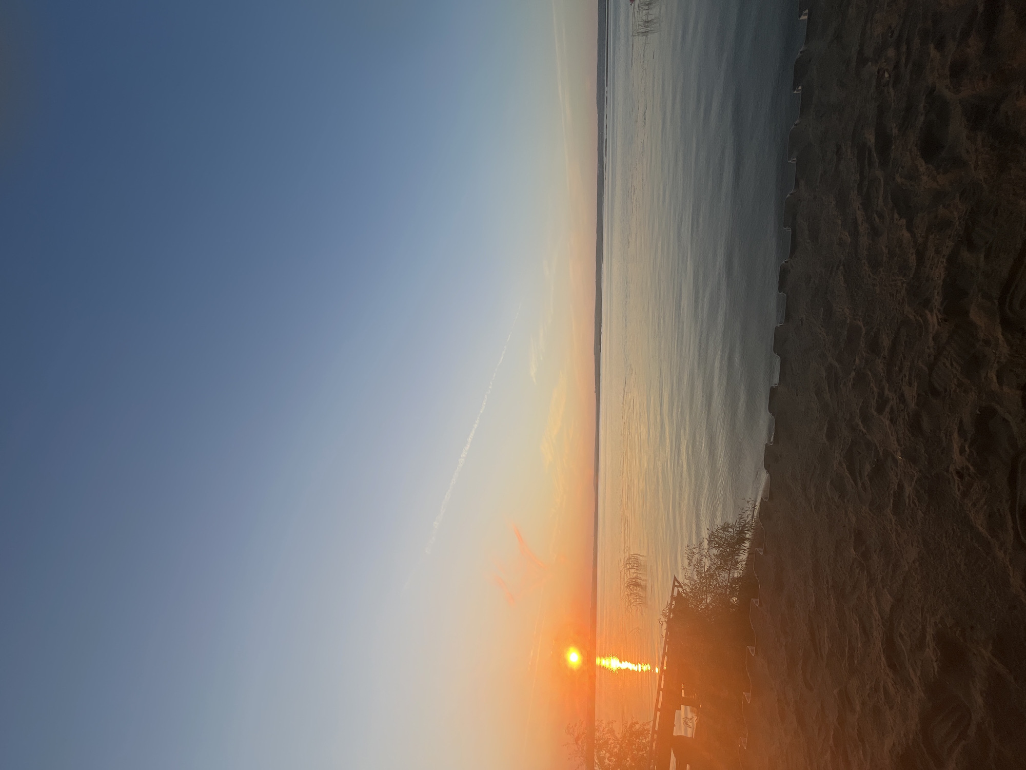 The image depicts a serene lake scene at sunset in Shumeika, Russia. The sun, a vibrant orange orb, is low on the horizon, casting a warm glow across the water. Its reflection creates a shimmering pathway on the lake's surface. The sky transitions from a pale orange near the sun to a soft blue at the top, with a faint contrail visible in the distance. The foreground consists of a sandy beach with visible footprints and a section of what appears to be a wooden structure or pier to the left, partially obscured by foliage. Reeds and other vegetation dot the shoreline in the mid-ground. The overall atmosphere is tranquil and peaceful. There are no people visible in the image.
