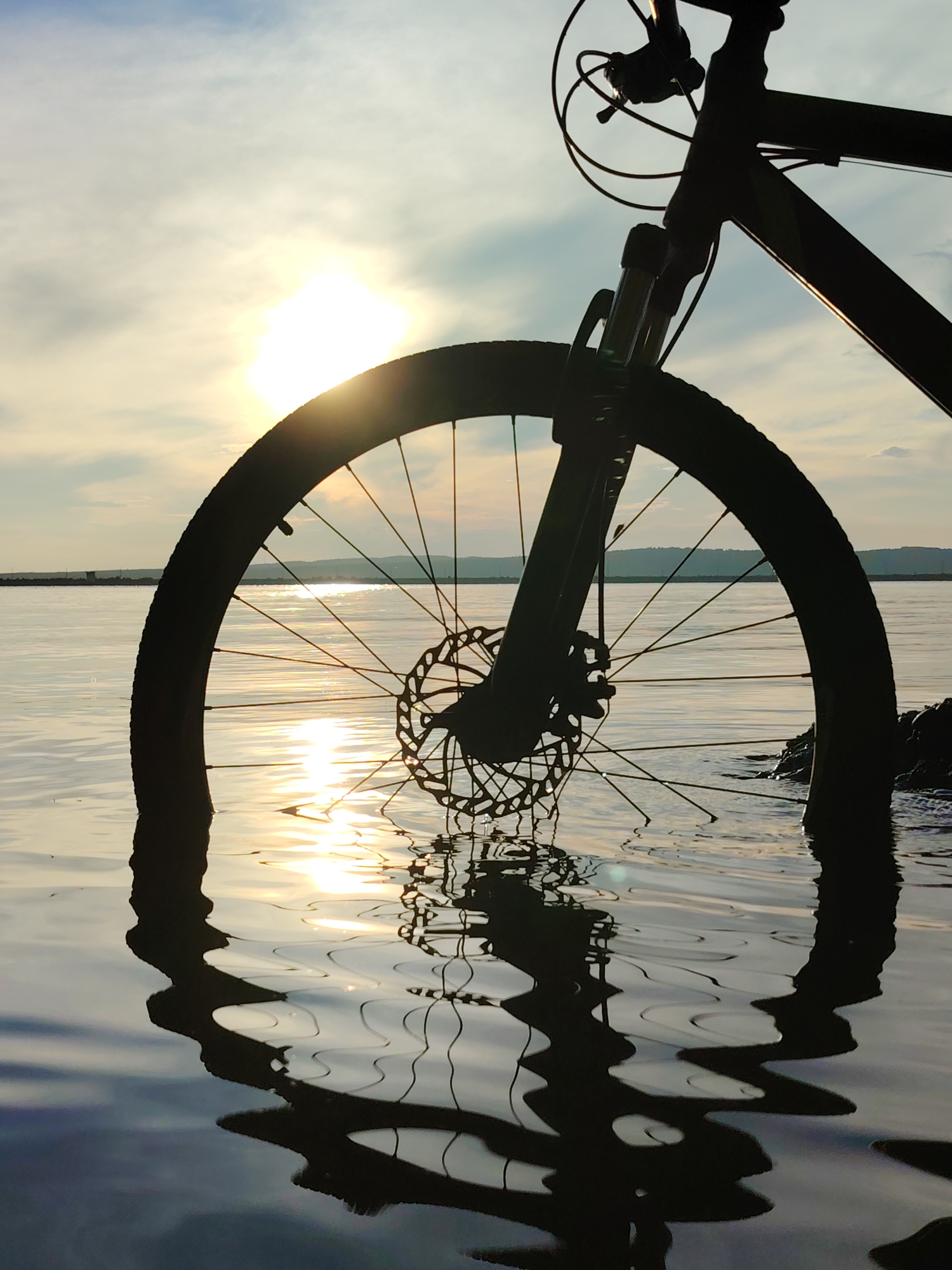 A bicycle is partially submerged in shallow water, with its front wheel and fork silhouetted against a bright sun and cloudy sky. The bicycle's frame and handlebars are also visible in silhouette. The water reflects the sun and the bicycle, creating distorted patterns. The background shows a calm body of water and a distant shoreline with hills. The scene appears to be set at sunset. The location is likely near the Kama River in Chaikovsky, Russia, given the characteristic landscape and lighting conditions often seen in this area during favorable weather. There are no people visible in the image.
