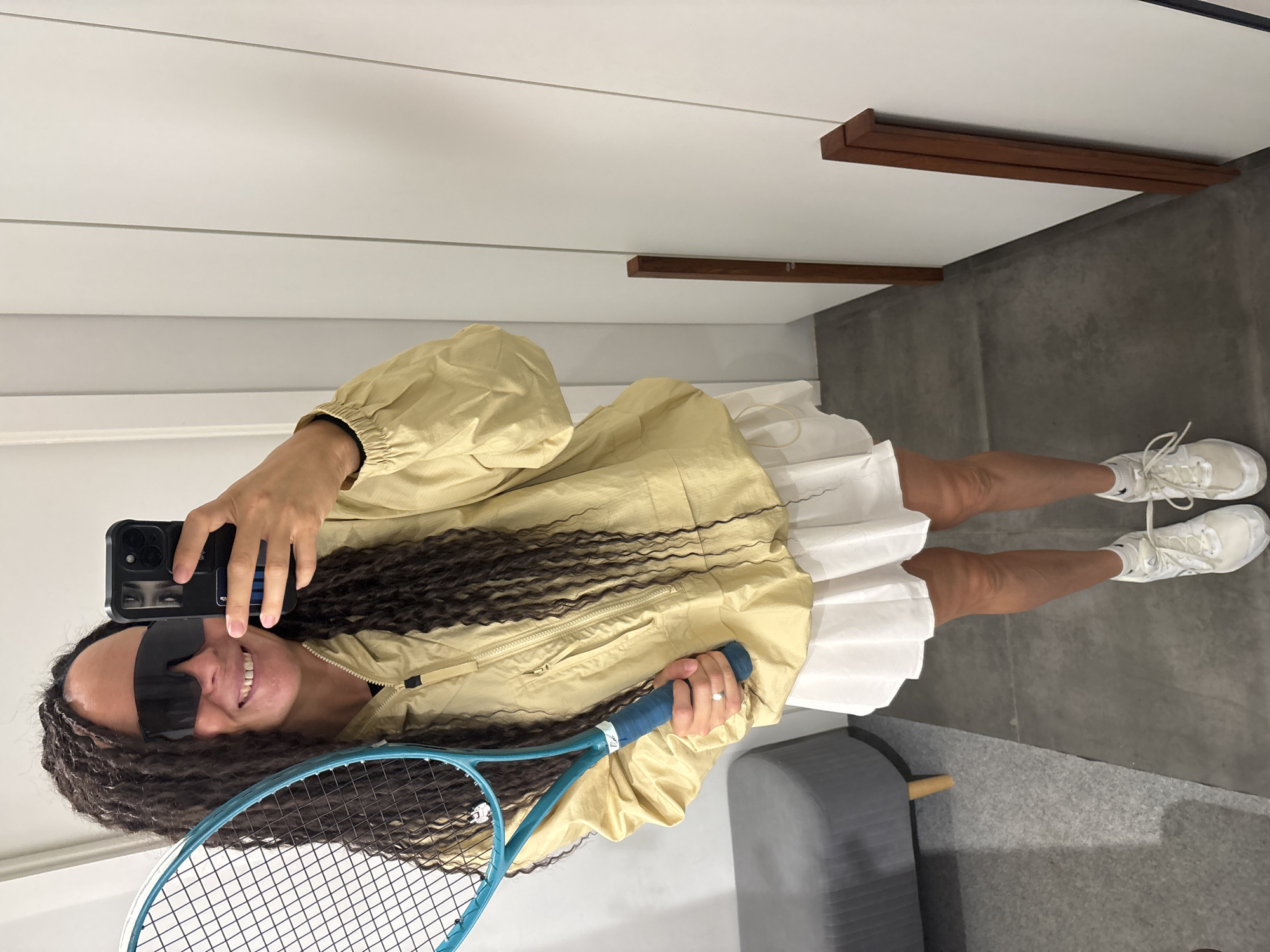 A woman with dreadlocks is taking a selfie in what appears to be a bathroom or dressing room. She's wearing a tan jacket over a white tennis skirt and white sneakers. She is holding a tennis racket in her left hand and a smartphone in her right hand. She's wearing sunglasses. The background consists of a white paneled wall with dark wood trim and a gray tiled floor. There is a gray ottoman with a wooden leg in the scene. There is no indication that this scene is located in Chaikovsky, Russia.