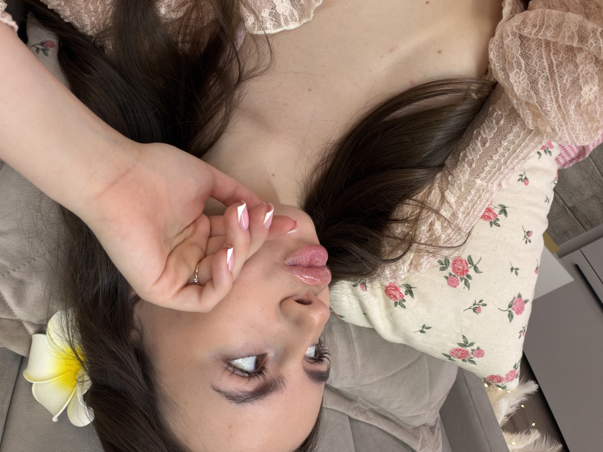 A young woman with long, dark brown hair lies on a gray upholstered surface, potentially a bed or couch. A white and yellow frangipani flower is tucked into her hair near her left ear. Her left hand is raised, obscuring part of her face, and she is wearing a gold ring on one of her fingers. She has long eyelashes and glossy lips. She is wearing a lace top with floral details and a pillow with a floral pattern. The image was taken indoors, possibly in a private residence. The location is Chaikovsky, Russia.