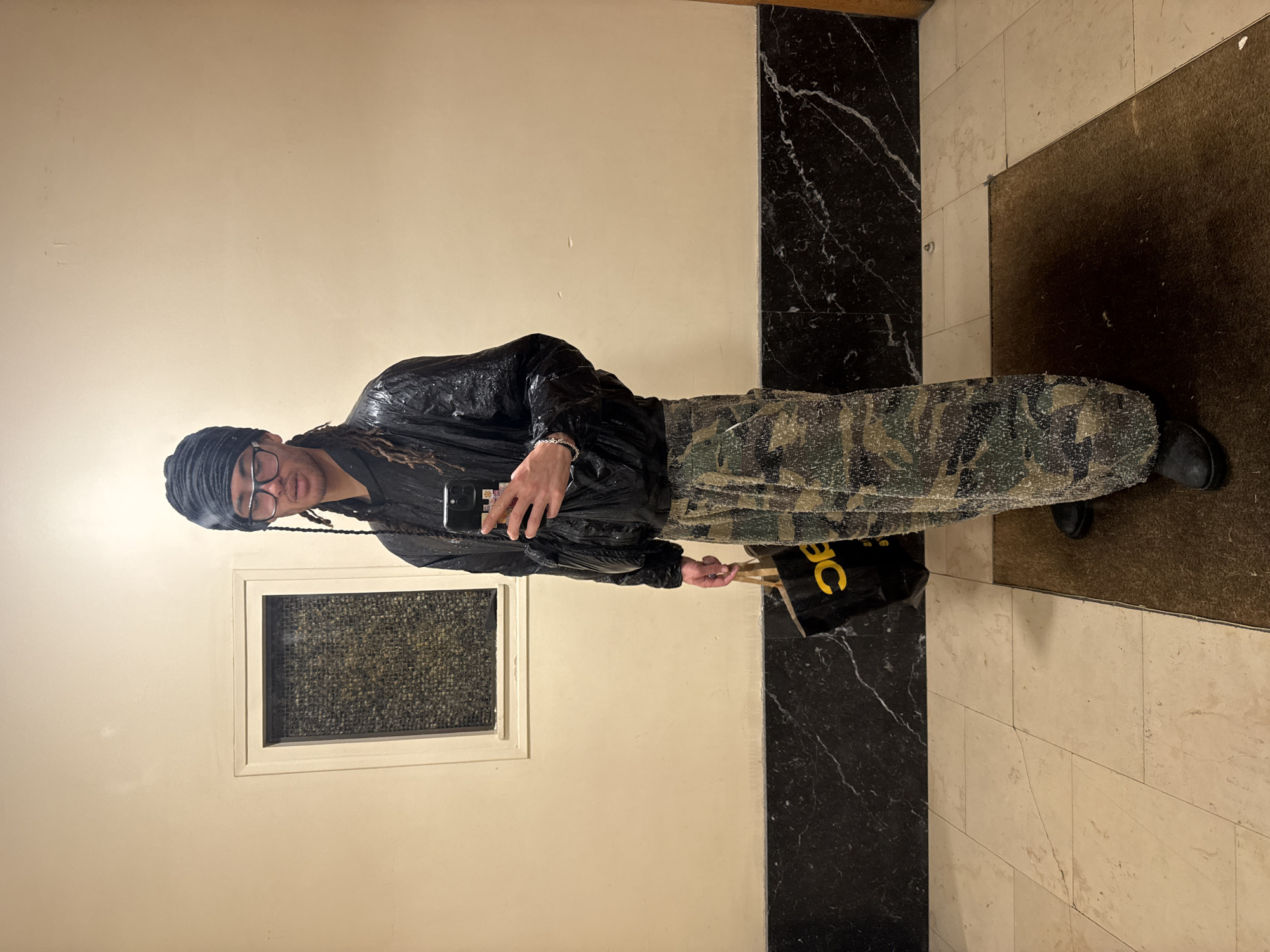 A person is standing in a room with beige walls and a black marble accent, holding a smartphone to take a selfie. The person is wearing a black head covering, glasses, a shiny black jacket, camouflage pants, and black shoes. The room has a small window and a shopping bag with yellow letters "BC." The location appears to be an interior space, potentially a building corridor. The city is not identifiable from the given image.