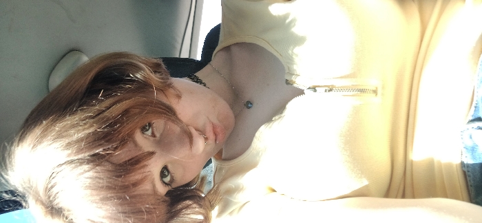 The image displays a head and torso shot of an individual, likely female, situated within what appears to be a vehicle interior. The person has short, light brown hair with bangs, fair skin, and light-colored eyes. A circular metal piercing is present in the nasal septum. A black choker necklace and a silver chain necklace with a small, dark pendant are worn around the neck. The individual is wearing a light yellow, ribbed top with a short zipper closure at the upper chest, and light blue denim material is visible at the bottom of the frame. The lips are pursed in a "duck face" expression, and the gaze is directed towards the camera. The background consists of an out-of-focus light-colored car headliner to the upper right and dark elements suggesting a seat or window frame to the left and behind. Bright light illuminates the subject from the left side, causing some overexposure. There is no discernible visual information within the image to specify a location in the city of Bistrice, Montenegro.