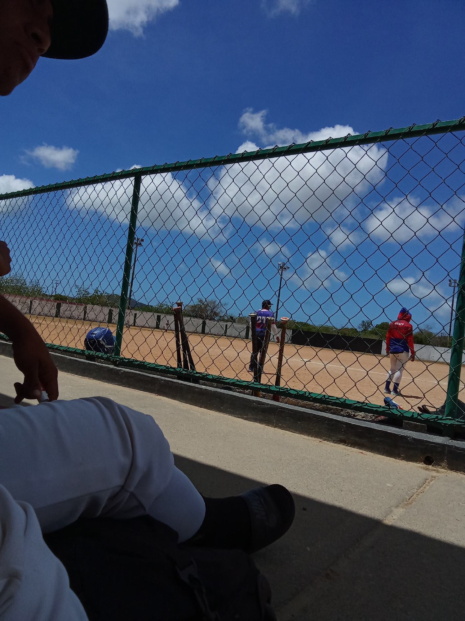 A person is observing a baseball field through a chain-link fence. Two individuals wearing baseball uniforms are on the field. One individual is wearing a purple and black uniform and another person is wearing a red and blue uniform with the word "STAFF" on it. There are stadium lights, a dirt field, and the sky is visible. The photograph may have been taken in the city of Juan Griego, Venezuela.