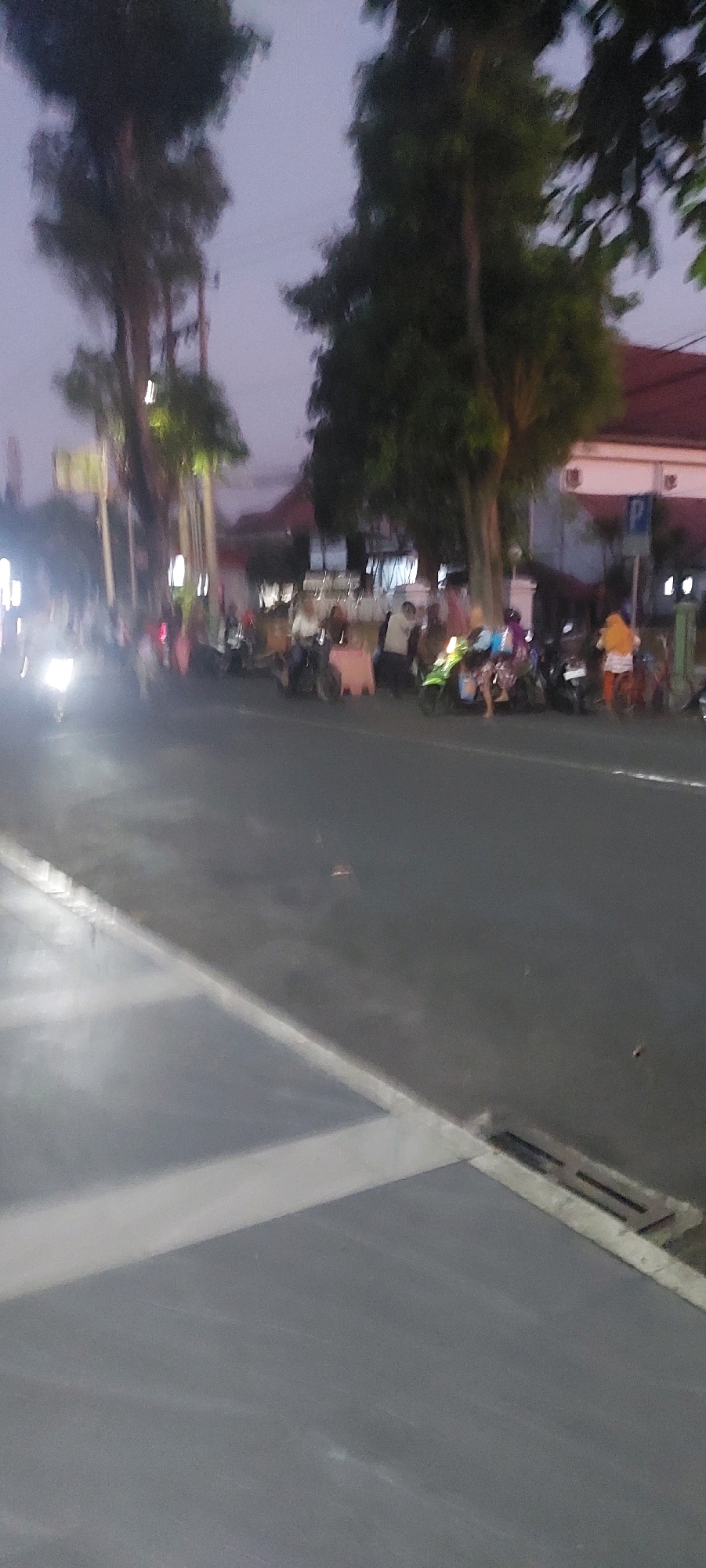 The image depicts a blurry, low-light street scene, likely in Lumajang, Indonesia, given the prompt. A paved road dominates the foreground, with a lighter-colored pedestrian walkway or sidewalk on the left. Several trees with dark foliage are visible along the right side of the road, obscuring some of the background buildings.
On the right side of the road, a group of people are gathered, some standing and some on or near parked motorcycles. The individuals appear to be casually dressed. One person on the far right wears a yellow top and is next to a red-and-white object. Another person is seated on a green motorcycle, wearing a blue top. Several other motorcycles are parked in a line.
In the background, a building with a red roof and white trim is visible, suggesting it might be a shop or establishment. A blue parking sign is also present near the building. On the far left of the image, a bright light from a vehicle is visible, indicating vehicular movement. The sky is a muted twilight hue, suggesting the photo was taken at dusk or dawn. The overall impression is of a street with some local activity taking place.